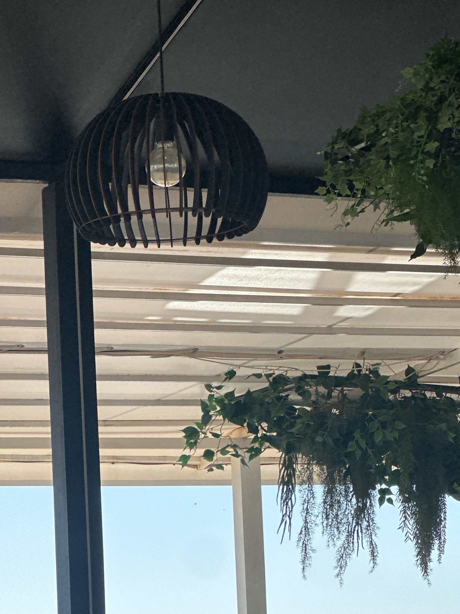 The image shows an outdoor seating area, likely a cafe or restaurant, with a covered roof supported by dark vertical beams. A decorative, spherical wooden pendant light fixture with an exposed bulb hangs from the ceiling. Several hanging planters with lush green foliage are suspended from the roof, adding a natural element to the scene. The background reveals a bright blue sky, suggesting it is daytime. The location is identified as Lermontovo, Russia.