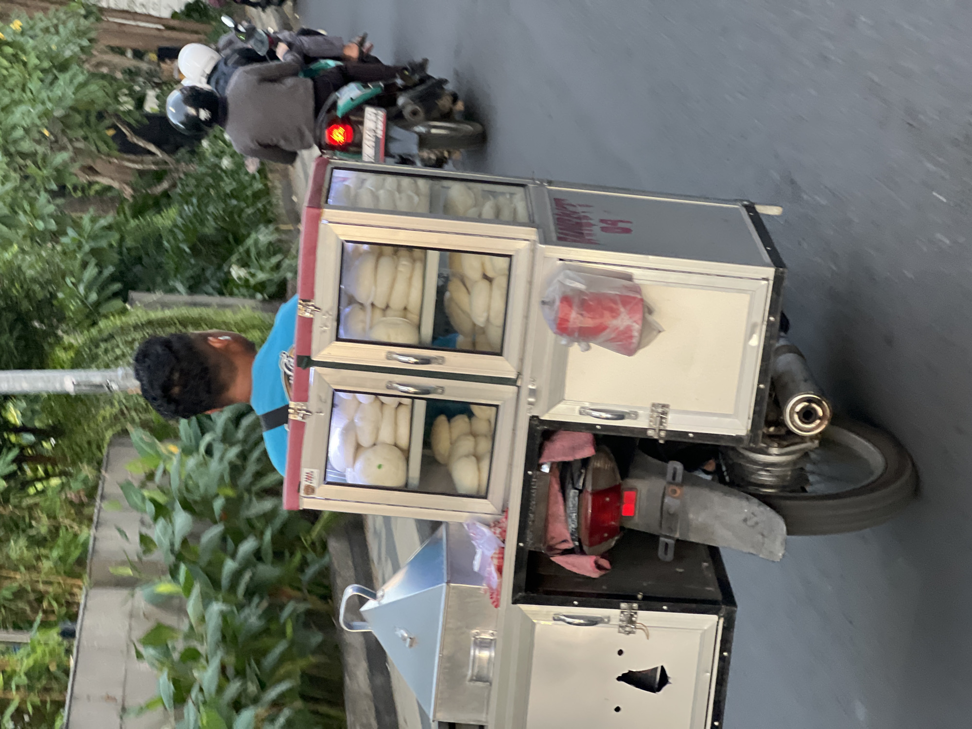 In Kuta, Indonesia, a food vendor navigates a street on a motorcycle. The vendor is seated on the motorcycle, with a food cart attached to the rear. The cart has a glass display showcasing steamed buns. The vendor is wearing a blue shirt. Other motorcyclists are visible in the background, also on motorcycles. The environment suggests a typical urban street scene with lush greenery bordering the road. The image is taken from a slightly elevated perspective, capturing the vendor and their mobile food setup in motion.