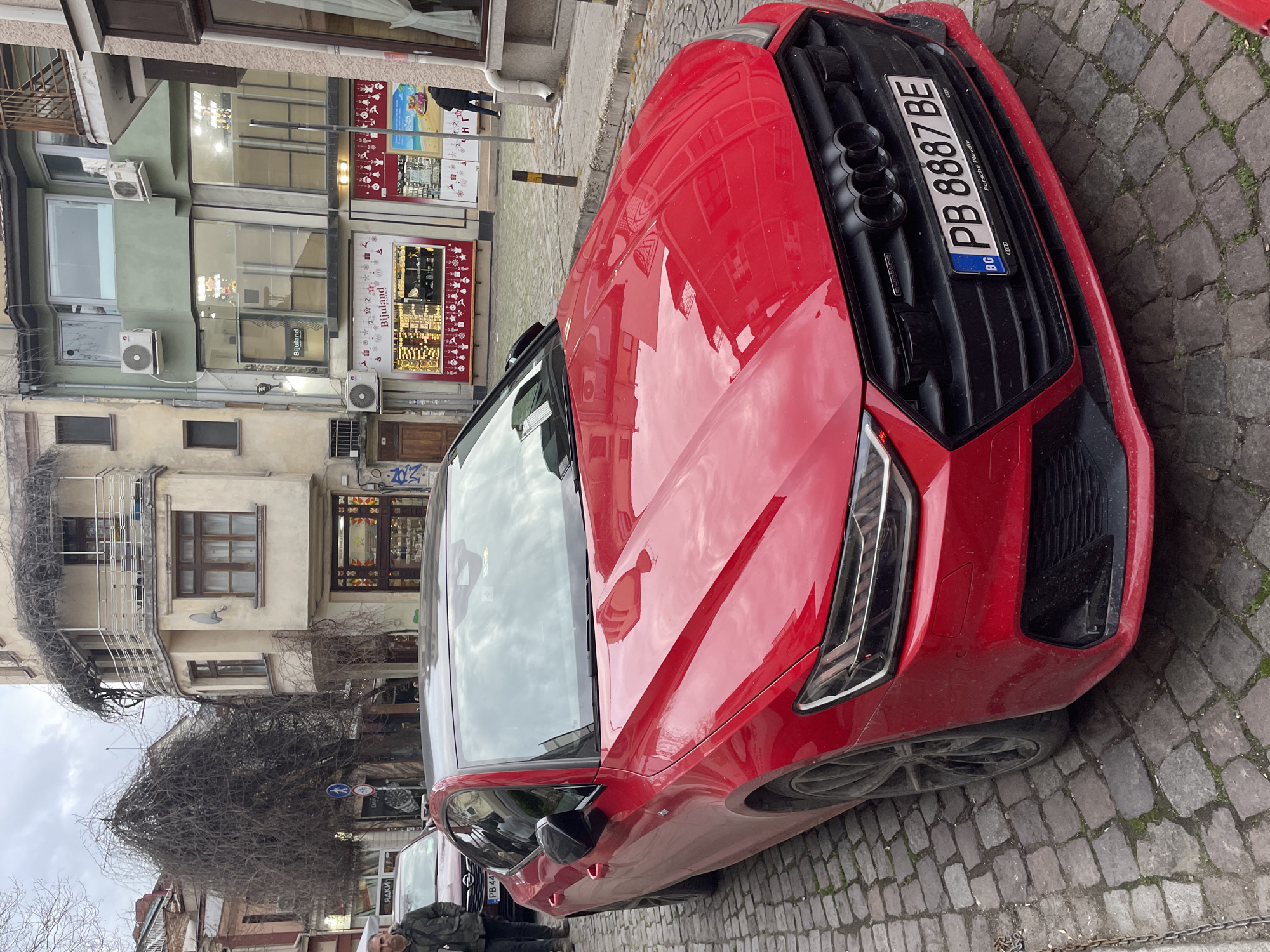 Here's a detailed description of the image:
**Subjects:** The primary subject is a red Audi car parked on a cobblestone street.
**Objects:** Besides the car, other visible objects include:
* Buildings with shops: "Bijuland" sign is visible.
* Air conditioning units on building facades.
* Street signs.
* Other parked cars.
* A person standing near another car.
* License plate on the Audi with number "PB 8887 BE". The license plate also has Porsche Plovdiv written on it and the BG abbreviation in the Bulgarian flag.
**Environment:** The setting is an urban area, evidenced by the buildings, shops, and paved street. The cobblestone street indicates a historical or older part of the city. The weather seems overcast.
**Location Clues:** The presence of the "Porsche Plovdiv" detail on the license plate and the Bulgarian license plate code "PB" suggest the location is in Plovdiv, Bulgaria.