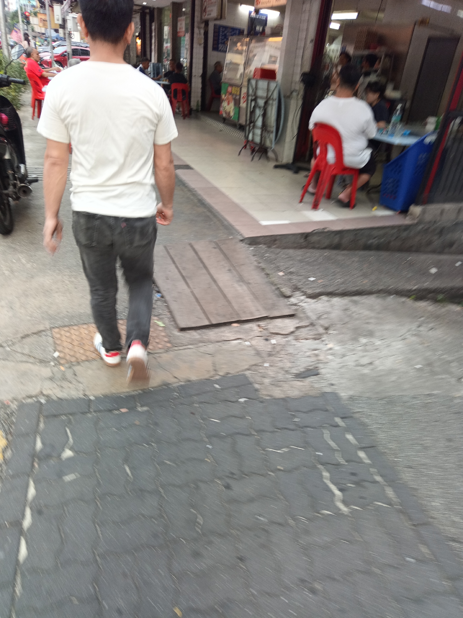A person walks away from the camera on a sidewalk in Kuala Lumpur, Malaysia. The person is wearing a white t-shirt and dark jeans. They are walking on a paved sidewalk with a motorcycle parked to their left. In the background, there are buildings with shops and people sitting at tables. The scene is blurry, suggesting motion blur from the camera or the subjects.