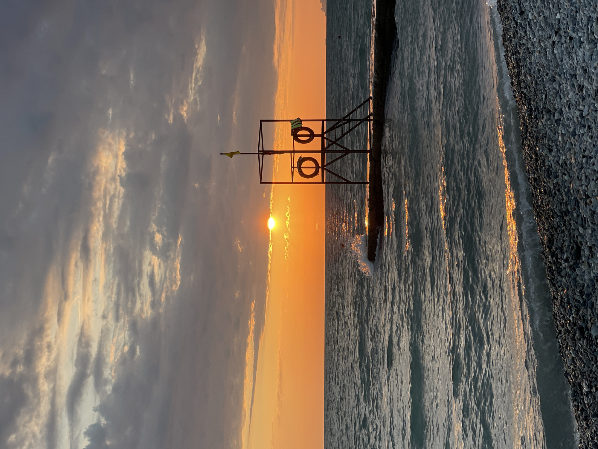 The image captures a tranquil sunset scene at a beach in Sochi, Russia. The sun, partially obscured by clouds, casts a warm orange glow across the sky and reflects on the rippling surface of the sea. In the foreground, a pebble beach lines the shore, with gentle waves lapping at its edge. A concrete pier extends into the water, supporting a lifeguard tower equipped with two lifebuoys and a yellow flag. The overall atmosphere is serene and picturesque, evoking a sense of calm as the day draws to a close.