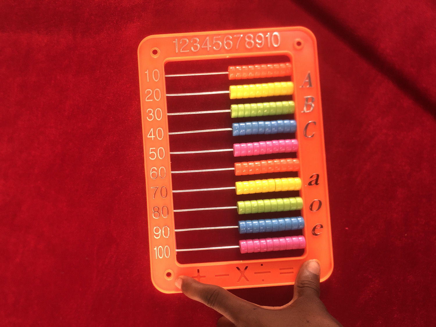 An overhead shot displays an orange plastic abacus, or counting frame, placed on a textured red fabric surface. A person's dark-skinned hand is visible in the bottom right corner, with an index finger pointing towards the mathematical symbols at the base of the abacus.
The abacus features ten horizontal rods, each holding ten colorful plastic beads. The beads are arranged in repeating color patterns: orange, yellow, green, blue, and pink. Along the top edge of the orange frame, the numbers "12345678910" are embossed. The left vertical side of the frame lists numbers "10, 20, 30, 40, 50, 60, 70, 80, 90, 100", indicating a hundreds counting aid. The right vertical side features embossed letters: "A, B, C" (capitalized) and "a, o, e" (lowercase), suggesting the toy also assists with alphabet recognition. At the bottom of the frame, mathematical operation symbols "+, -, ×, ÷, =" are visible.
The scene appears to be indoors, likely in a learning or play environment, with good, even lighting. The hand's position suggests engagement with the educational toy, possibly for learning basic arithmetic or counting.