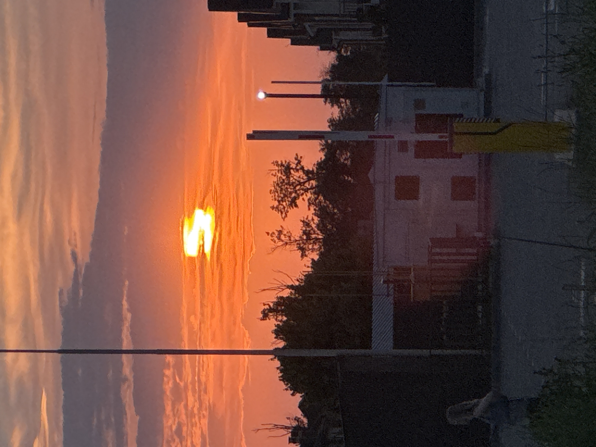 A high-angle, medium shot captures a dramatic sunset over an urban industrial area. The sky is ablaze with vibrant orange and red hues, with scattered clouds adding texture. The sun itself is partially obscured by clouds, creating a bright, diffused glow. In the foreground, a tall, slender pole stands vertically on the left, its top disappearing out of frame. To the right, a series of buildings with dark facades and pointed rooftops create a jagged skyline. A white building with dark square windows is visible in the middle ground. Two white barrier gates with red accents are positioned in front of this building. A bright yellow pole with a ribbed texture stands near the right barrier gate. In the far left of the frame, a person is partially visible, facing away from the camera, with their arms outstretched. The ground is paved, with some grass and foliage growing in the lower portion of the image. The scene appears to be an outdoor industrial or security checkpoint area at dusk.
