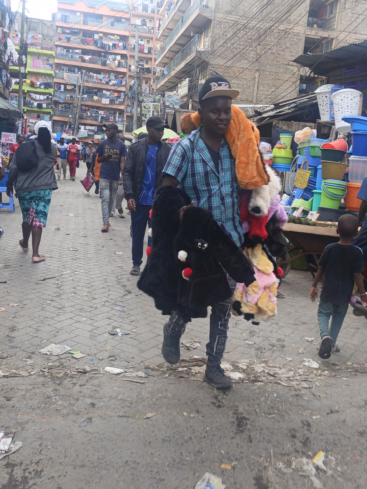 The image captures a bustling street scene in Nairobi, Kenya. A man wearing a plaid shirt, ripped jeans, and a baseball cap walks towards the viewer, carrying a large assortment of plush toys and blankets over his shoulder and in his arms. He appears to be a vendor. To his right, a young boy in a dark t-shirt and light blue pants walks in the same direction.
Behind the vendor, other pedestrians populate the street. Buildings of varying heights and colors line the street, with clotheslines strung between them. A collection of plastic buckets and containers is visible near the edge of the frame, possibly another vendor's wares. The ground is littered with trash and debris. Overhead, a dense network of electrical wires crisscrosses the sky.