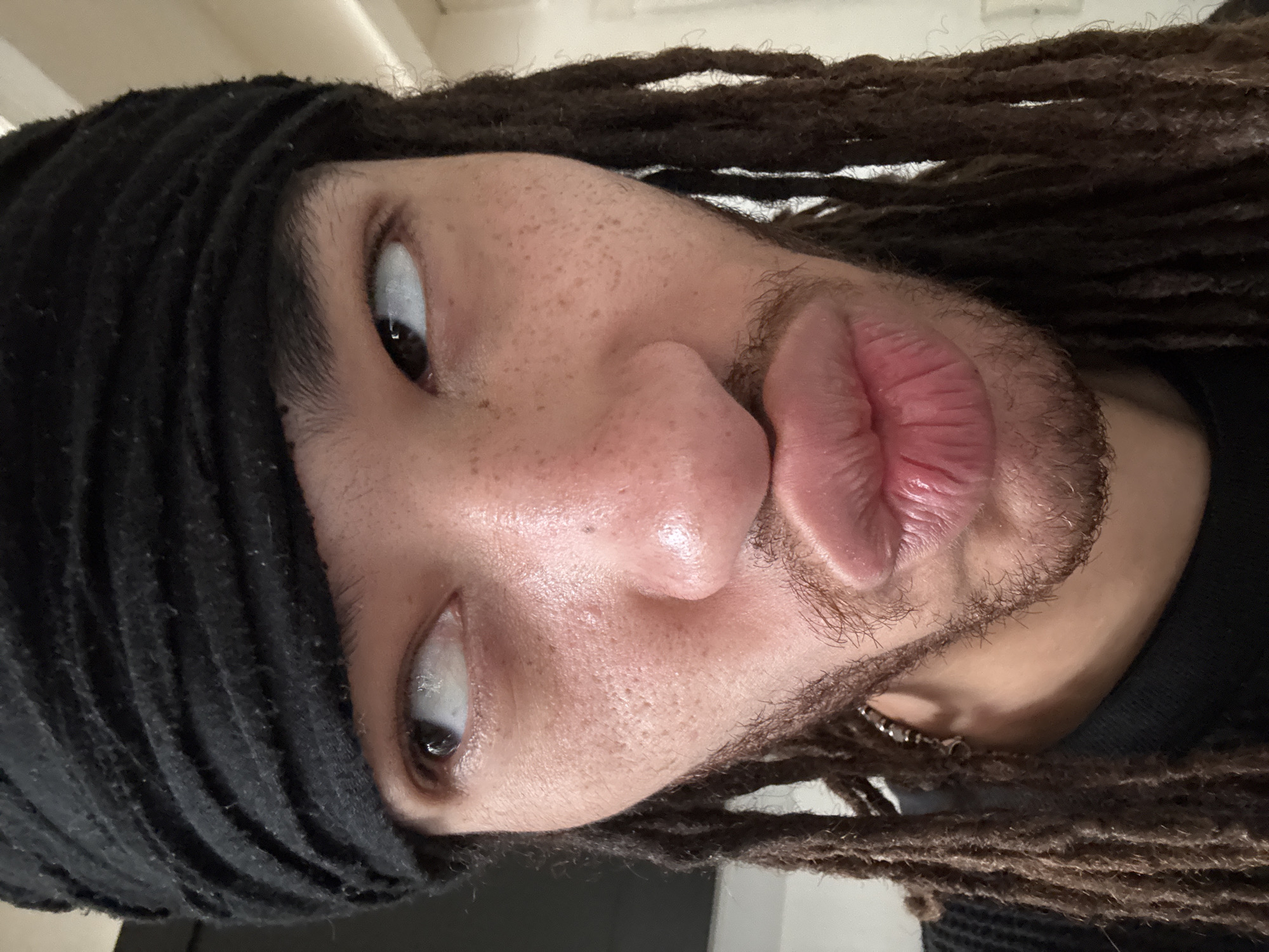 A close-up headshot photograph depicts a person with light brown skin and a short, dark brown beard, making a pouting expression. They have light brown eyes and freckles on their nose and cheeks. A black, ribbed beanie-like fabric covers part of their head, and dark brown dreadlocks frame their face. An earring is visible near the jawline. The background is a blurred off-white. The photograph appears to be taken indoors under soft, diffused light. There is no contextual information to suggest this photo was taken in Paris, France.