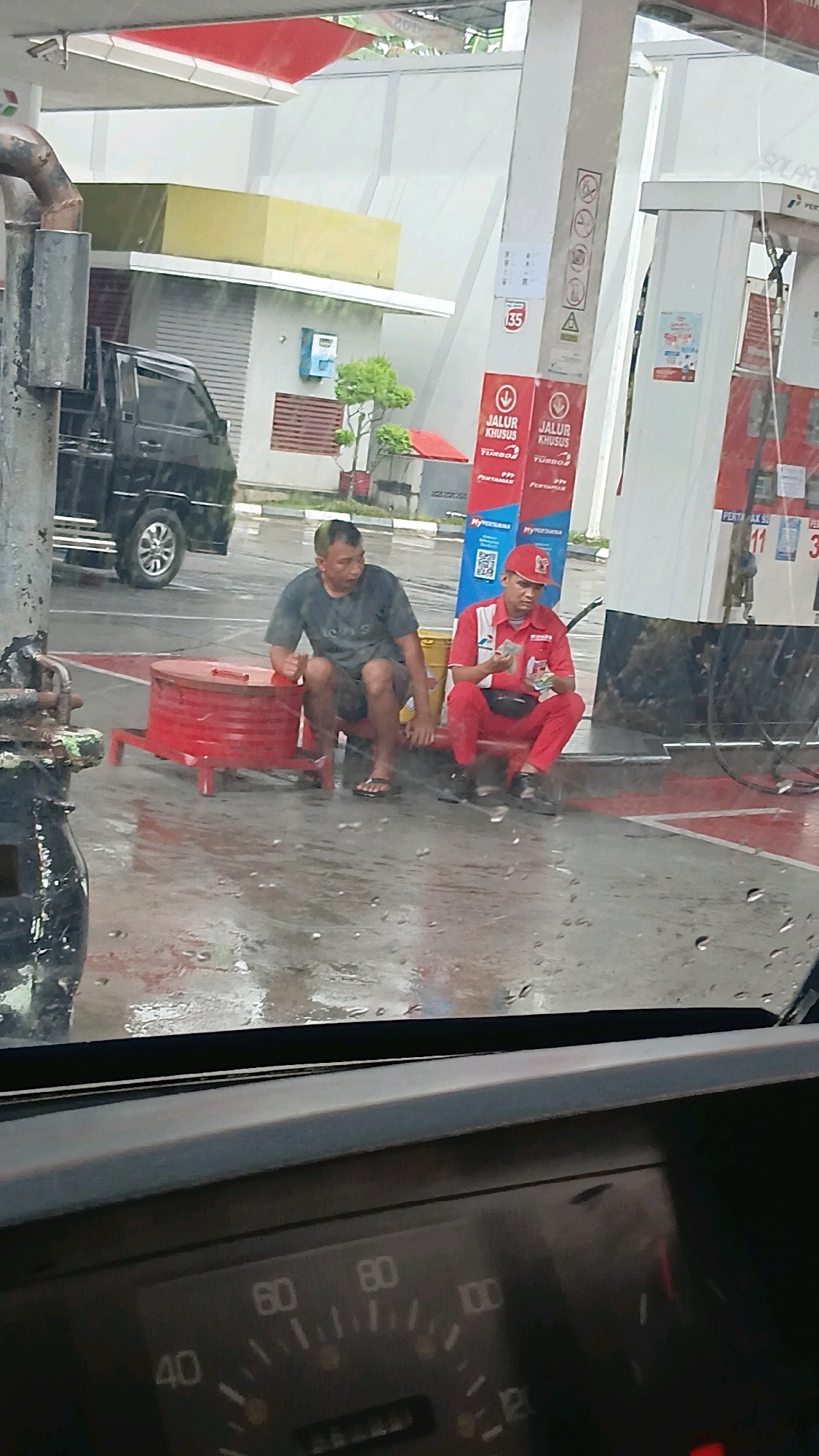 The image shows a rainy day at a Pertamina gas station in Batusangkar, Indonesia. Two men are sitting on red seats near a fuel pump. One man, wearing a dark t-shirt and shorts, is looking to his left. The other man, wearing a red uniform and cap, is holding and counting cash. A black pickup truck is parked in the background. The ground is wet and reflects the surrounding structures. The image is taken from inside a vehicle, with a portion of the dashboard and speedometer visible in the foreground.