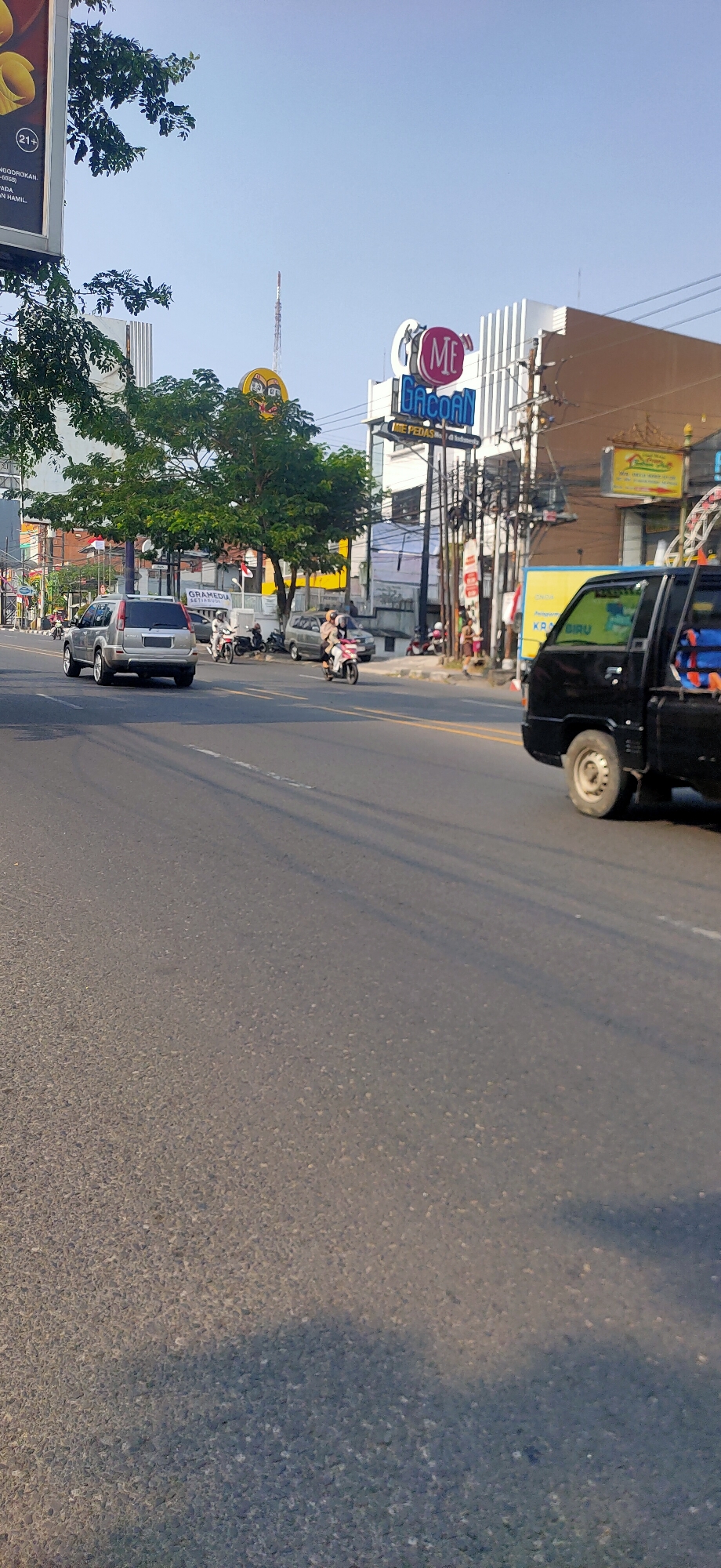 The image depicts a street scene in Semarang, Indonesia, with a focus on the road and surrounding buildings. In the foreground, the asphalt road shows tire marks and is partially covered by shadows. A black pickup truck is visible on the right side of the road. Further down the street, a silver SUV is traveling, and several motorcycles are present. Buildings line the street, including a structure with a prominent "ME GACOAN" sign. Trees and utility poles are also visible. The sky is clear blue.