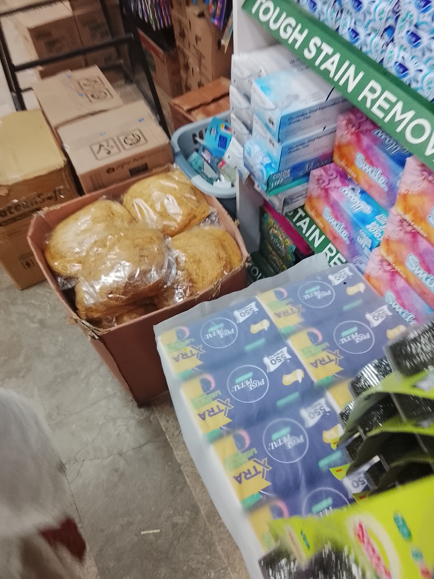 The image shows a retail setting, possibly a small store or warehouse in Gulgasht Colony, Pakistan. Items visible are household cleaning products, including stain removers, disposable gloves, and Rose Petal-branded paper products, stacked next to boxes of what appears to be wrapped food items. The products are arranged in cardboard boxes and plastic crates. The floor appears to be tiled or made of concrete. A partial view of a person wearing a white garment is visible in the lower-left corner.