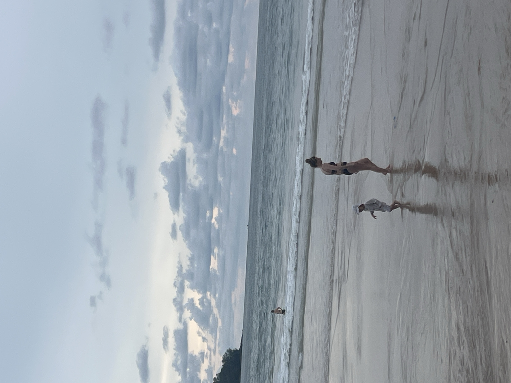 A group of people are on a beach in Choeng Thale, Thailand. In the foreground, a woman in a black bikini walks along the wet sand, and a child in a light-colored outfit walks in the shallow water. Further out in the ocean, a person stands in the water. The sky is overcast with clouds.