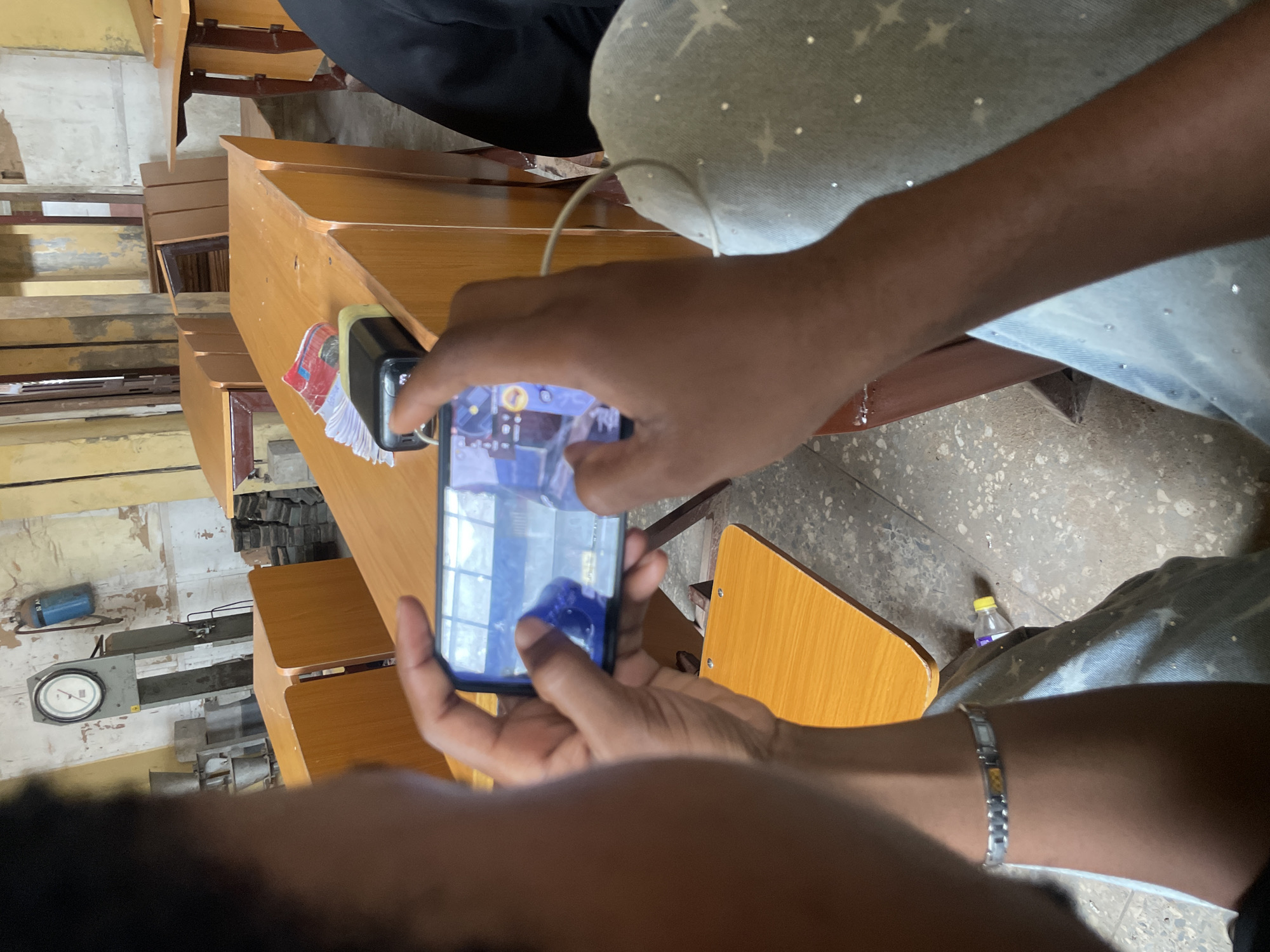 In a classroom setting in Ihiagwa, Nigeria, a person is captured from behind, holding a smartphone horizontally to play a game. The person is wearing a silver bracelet on their left wrist and light blue jeans adorned with small, sparkling star-like embellishments. The phone's screen displays a colorful game interface. In the background, several wooden desks and chairs are arranged, with some appearing to be stacked. On one of the desks, a black power bank and a stack of paper money are visible. The environment suggests a functional but somewhat utilitarian space, with concrete flooring and walls that show signs of wear. A blue gas cylinder hangs on the wall in the upper left corner, and a scale is mounted nearby.