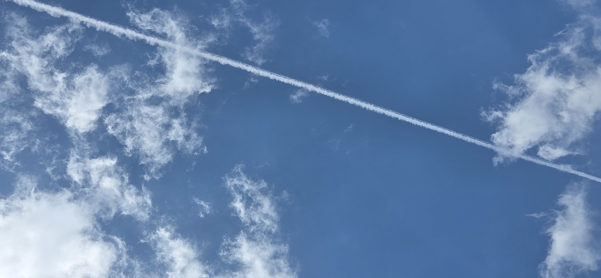 The image shows a clear blue sky with scattered white clouds. A prominent contrail, formed by an airplane, stretches diagonally across the frame from the bottom left to the upper right. The contrail is a bright white line against the blue sky. The clouds are wispy and appear to be cirrus clouds. There are no people, buildings, or any other objects visible in the image. This image is a photograph of the sky and does not depict any specific location in Karlsruhe, Germany.