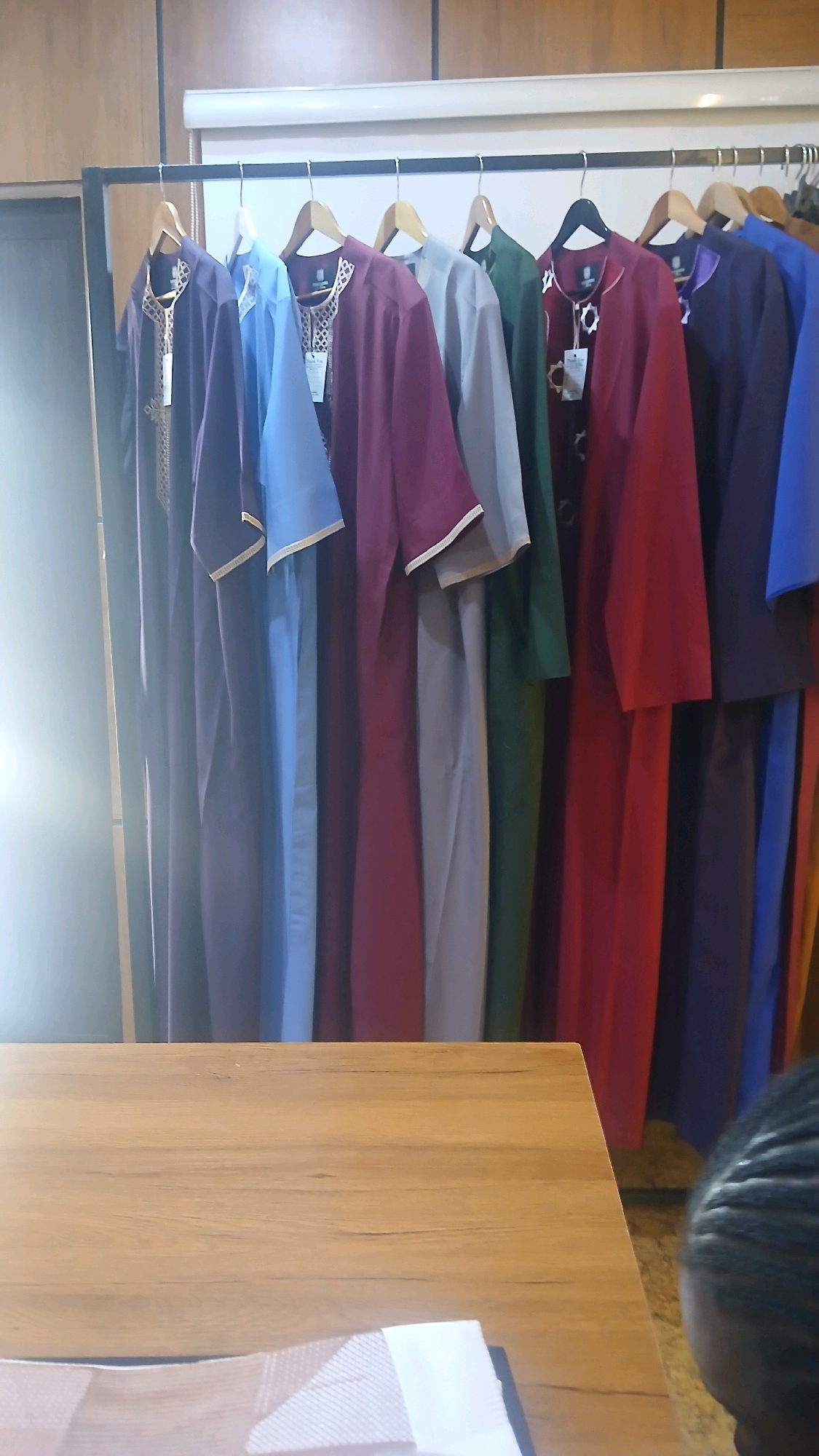 The image shows a rack of colorful men's traditional clothing, specifically jalabiyas, displayed in a store. The jalabiyas are in various shades of purple, blue, maroon, gray, and green, with some featuring gold or white embroidery around the neckline and cuffs. They are hung on wooden hangers from a metal rack. In the foreground, a portion of a wooden table and a person's braided hair are visible, suggesting a customer browsing in the store. The background features wooden paneling on the walls. The store appears to be located in Gura Top, Nigeria, a commercial area known for its clothing vendors.