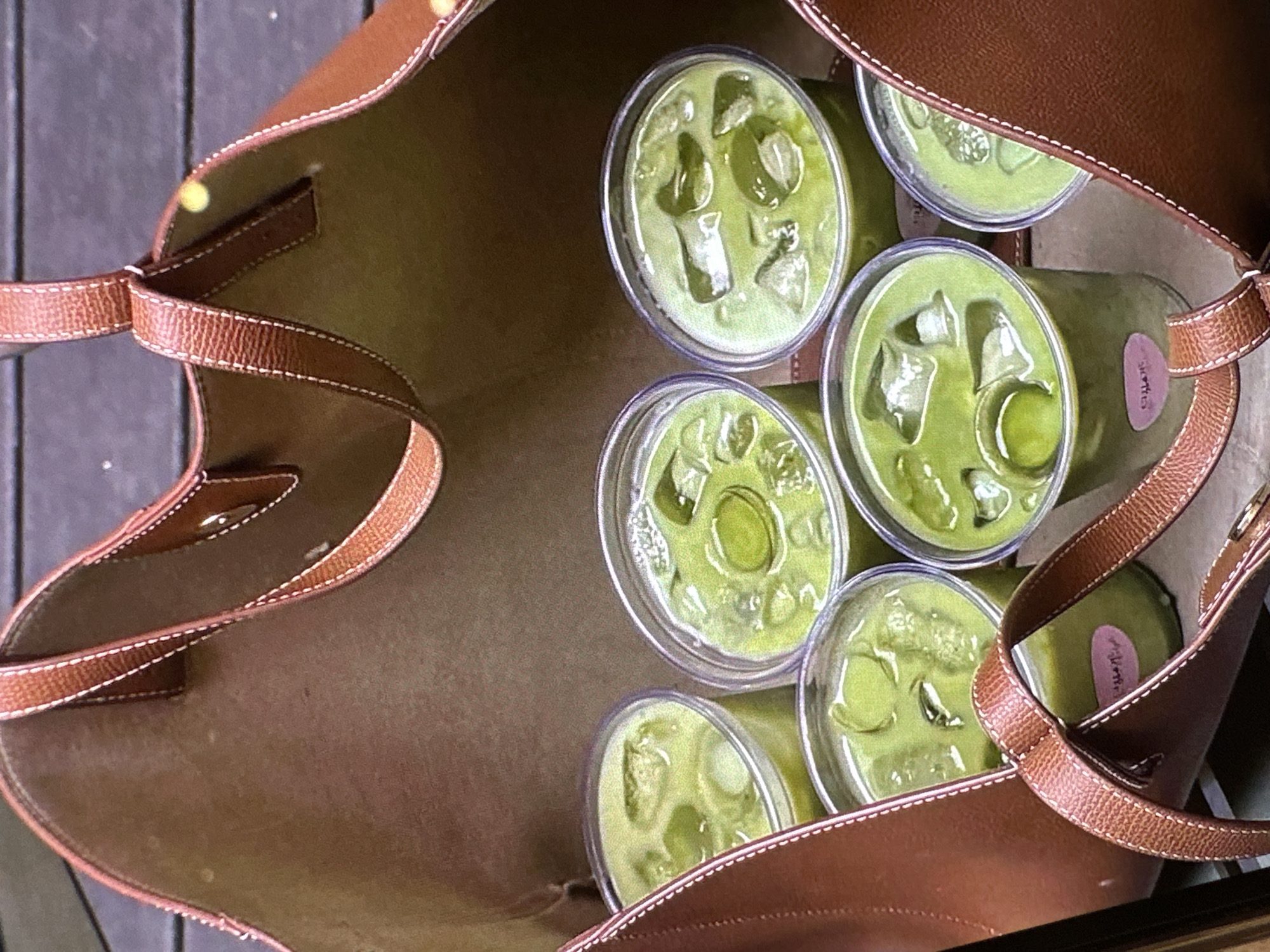 The image shows a brown leather tote bag filled with six clear plastic cups containing a light green liquid and ice, likely matcha lattes. The bag's interior is a light brown color. The cups are arranged closely together inside the bag. The bag is presumably on a wooden surface, based on the planks seen in the background. The location is unknown as Yerevan, Armenia, cannot be confirmed from the visual evidence.