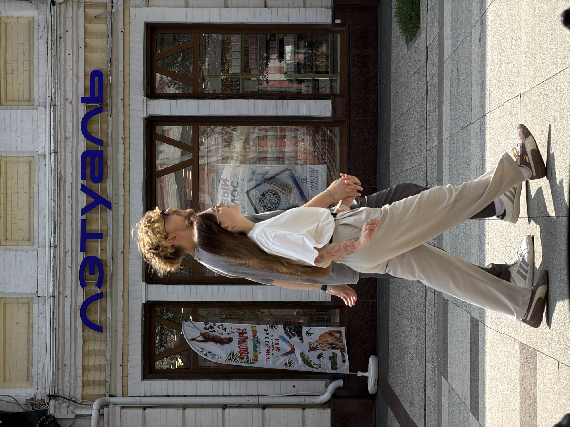 The image captures a daytime scene on a sidewalk in front of a building. Two individuals, a man and a woman, are walking side-by-side, holding hands. The man has curly blonde hair and wears glasses and a gray t-shirt. The woman has long brown hair, wears a white t-shirt, and light-colored pants, and has visible tattoos on her arm. They are walking on a paved sidewalk with a checkered pattern.
Behind them, the building features a white brick facade with yellow accents and large windows. A prominent blue sign with Cyrillic lettering is mounted above the windows. To the left, a tall banner-style flag displays images of various animals, including an orangutan, a lizard, a bird, a panther, and a spider, along with text in Russian. The windows of the building appear to be shop windows, with displays of merchandise visible inside, including what looks like cosmetics and branded goods like "GUCCI." The overall environment suggests an urban setting, likely a commercial street. The text on the blue sign appears to read "АКТУАЛЬ" which translates to "ACTUAL" in English. The banner also contains text, with "ЗООПАРК" (Zoo Park) being clearly visible. Given the presence of Cyrillic script and the likely identification of the store as "АКТУАЛЬ", this scene is located in Saratov, Russia.