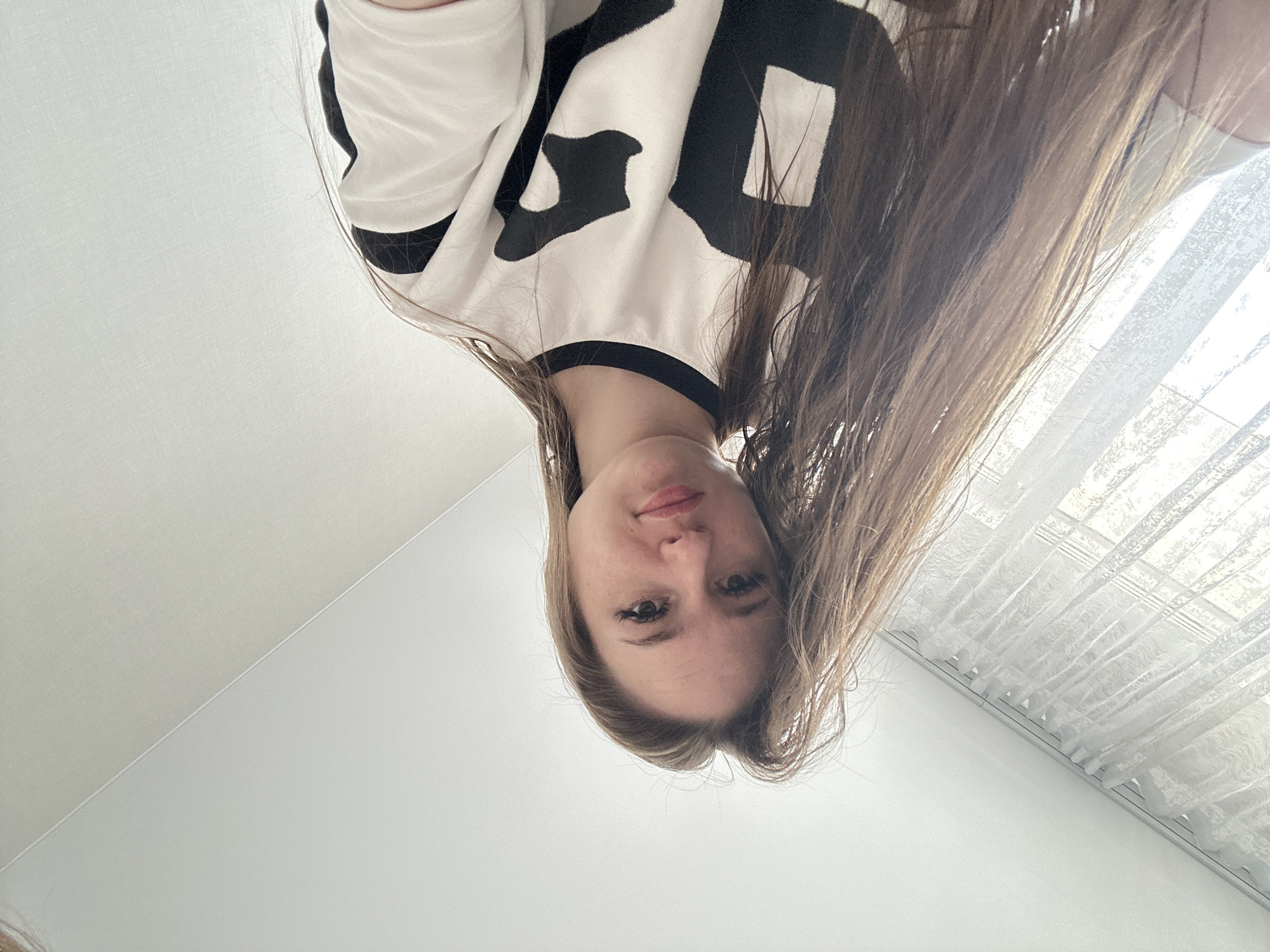 A young woman with long, light brown hair is the central subject of the image, depicted from an unusual low-angle perspective with her head inverted in the frame. She looks directly at the camera with a neutral to slightly pleasant expression and a subtle smile. Her eyes are open and focused. She is wearing a white long-sleeved top featuring prominent, large, black, blocky, stylized patterns across the front. These patterns could be abstract shapes or highly stylized letters.
The setting appears to be indoors, with plain white walls or ceiling dominating the background, giving a clean and minimalist feel. To the right, a window is visible, covered by white sheer curtains, allowing bright daylight to filter into the room. The lighting suggests it is daytime. The exact location is not specified beyond being a modern interior room. No specific text is clearly decipherable on her clothing, only the aforementioned stylized patterns.