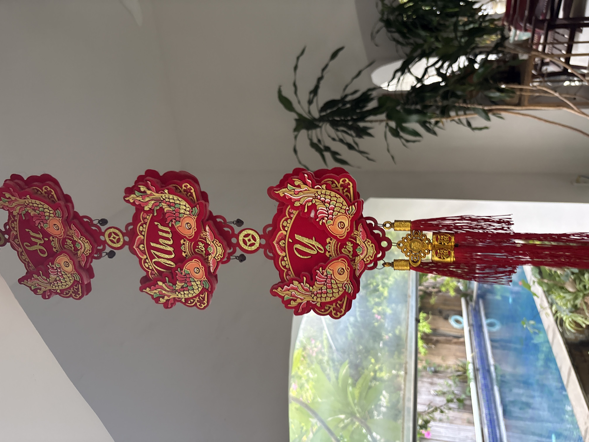 Three red and gold ornamental fish decorations are suspended from the ceiling. The decorations are layered and feature intricate patterns and Chinese characters. The background shows a blurred view of a swimming pool, lush greenery, and a potted plant. The scene is captured from an interior perspective, looking out towards an outdoor area. The location is not specified in the image or provided information.