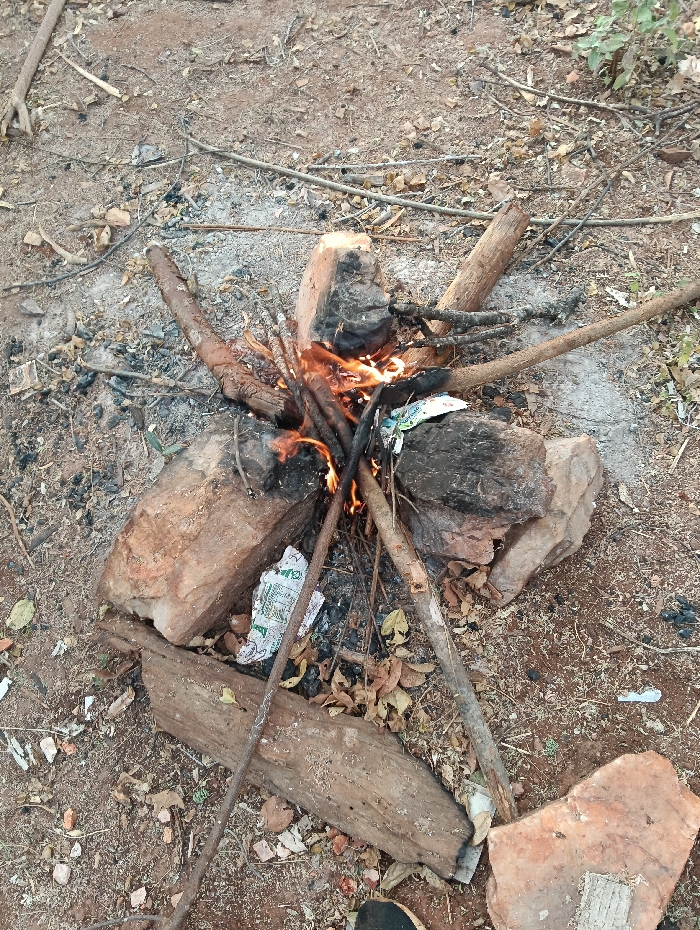 A small, active bonfire is observed at ground level, situated on dry, reddish-brown earth. The fire is contained by a perimeter of large, irregular rocks. Various sizes of wooden sticks and logs serve as fuel, with several pieces visibly charred and actively burning, producing bright orange flames at the center. Scattered around the fire on the soil are dry leaves, small branches, and ash. Two crumpled white wrappers, one with green markings, are visible near the fire's base. No individuals are present in the frame. The scene is located outdoors in Kyamatu, Kenya.