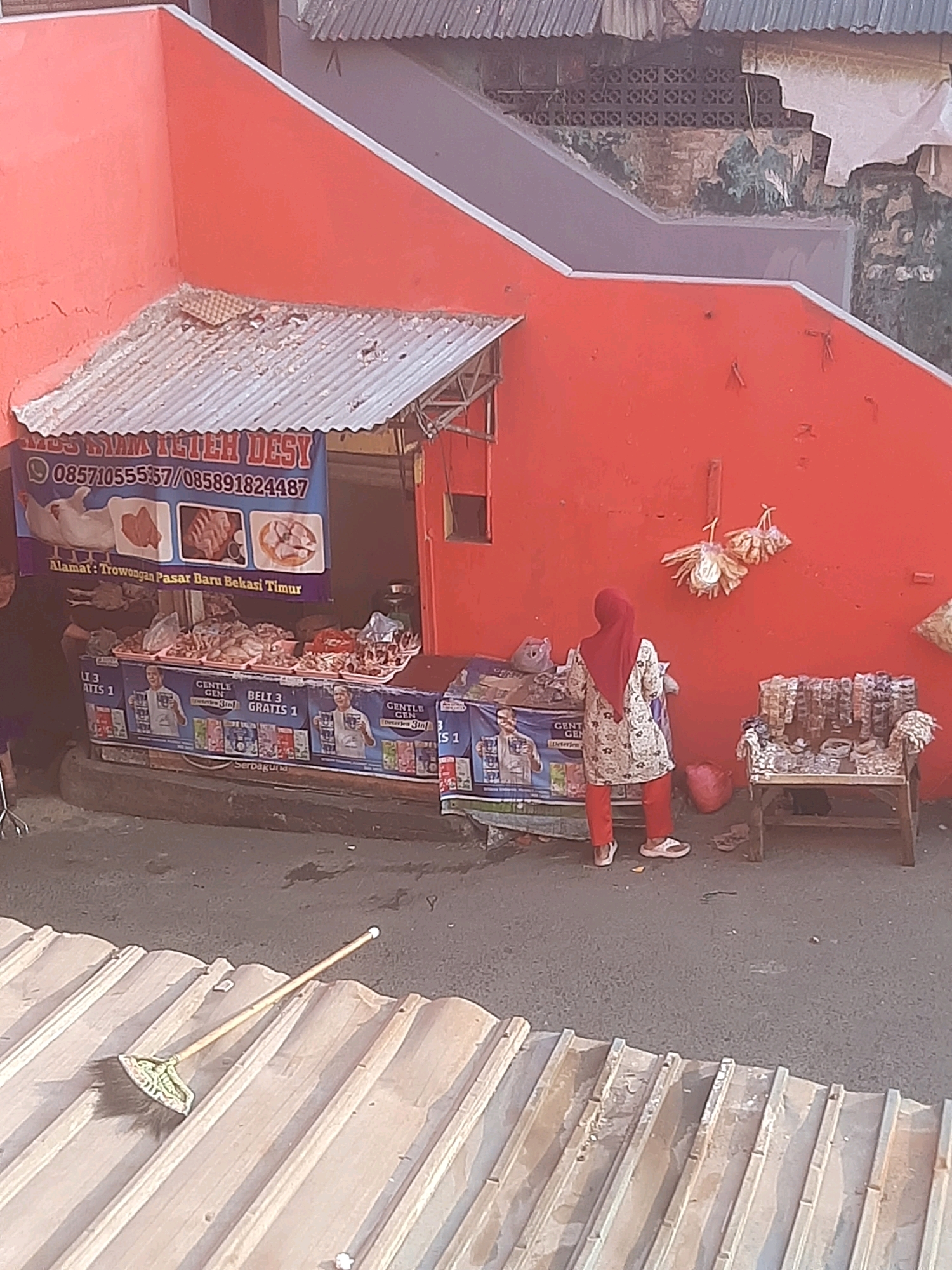 The image shows a street-level view of a small market stall in Bekasi, Indonesia. The stall is decorated with a large blue banner featuring images of chicken and text, including "MOS AYAM TETEH DESY" and phone numbers. Below the banner, there are trays filled with raw meat, likely chicken, displayed for sale. A woman wearing a red headscarf and a patterned shirt is standing in front of the stall, facing away from the camera. She is wearing red pants and white sandals. To the right of the stall, there is a wooden bench with various items displayed on it, possibly snacks or small goods. In the foreground, a broom rests on a corrugated metal roof. The stall itself is constructed with a metal roof and appears to be part of a larger building with an orange facade. The overall scene depicts a typical market setting in an urban area.