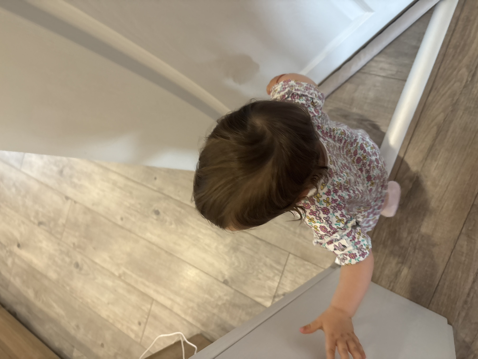A young child with brown hair, wearing a floral print outfit and light pink shoes, is standing on a wooden floor in what appears to be a doorway. The child is reaching out with their left hand towards a light gray surface on the left side of the frame. A white door or wall is visible on the right. The lighting suggests an indoor environment. There is no information available to specify the location in the city of Strelcha, Bulgaria.