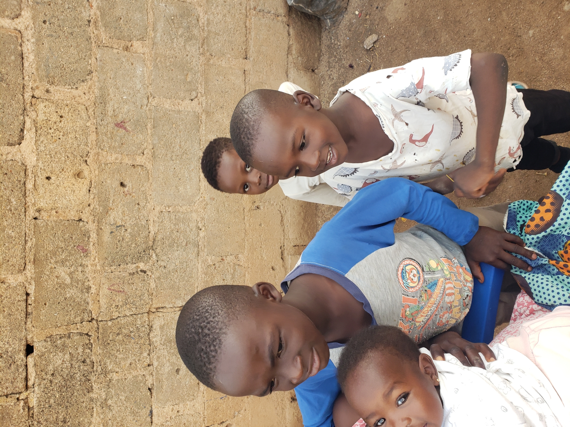 In the city of Jos, Nigeria, four young children are gathered in front of a textured, beige brick wall. A young child, possibly a girl, is being held by an older child, her face turned towards the camera with wide, curious eyes. The older child, a boy wearing a blue and gray t-shirt with colorful graphics, looks away from the camera with a thoughtful expression. To the right of this pair, another boy, the youngest of the three visible older children, is smiling and looking towards his left, wearing a white t-shirt adorned with a pattern of stylized birds. Behind him, a fourth child, partially obscured, also appears to be looking towards the left. The ground beneath them is sandy and uneven, with scattered debris. The overall scene captures a moment of childhood interaction in a simple, outdoor setting.