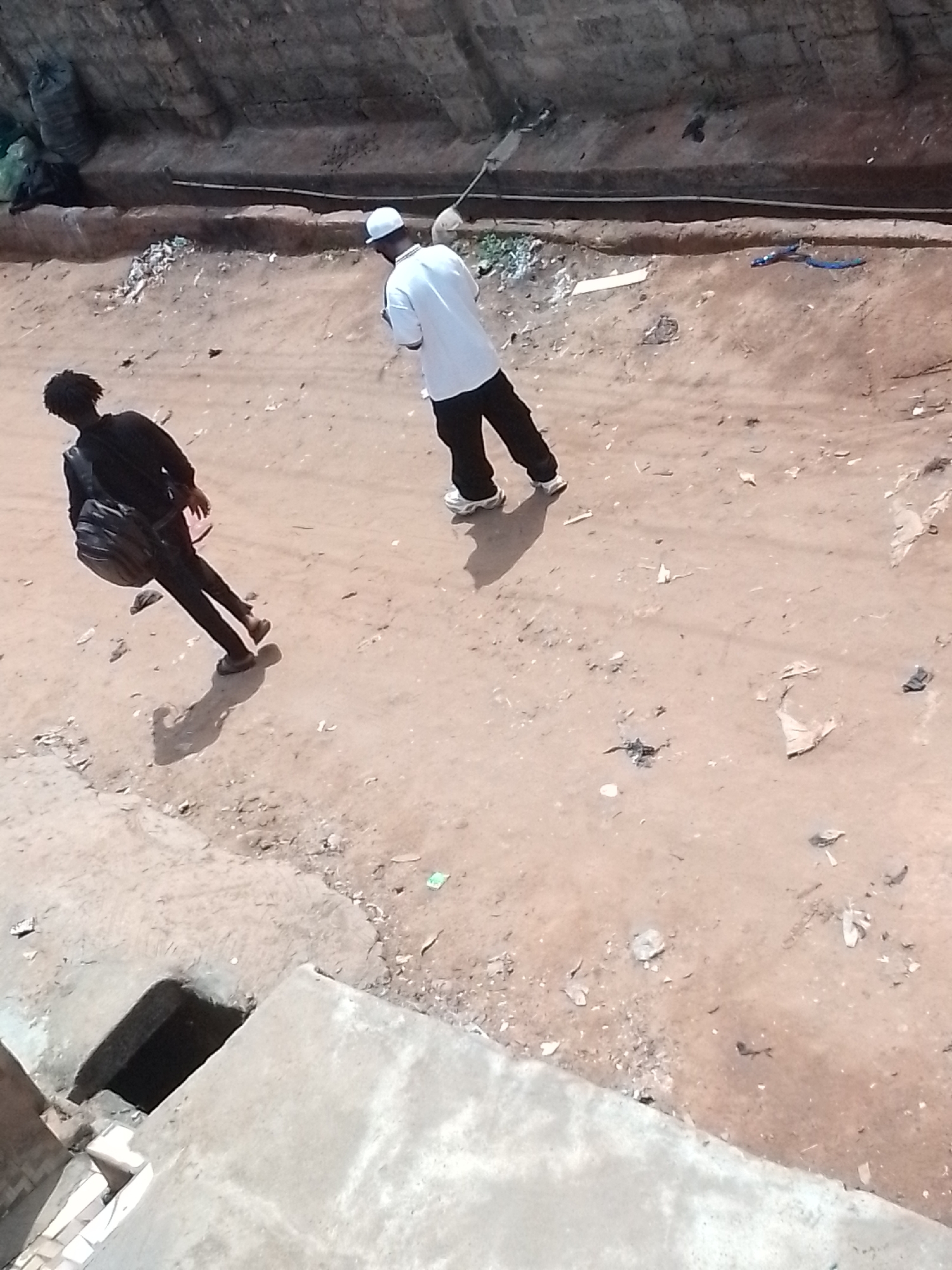 The image shows an outdoor scene in what appears to be an urban area. Two individuals are visible. One is wearing a white t-shirt, black pants, and a white cap, while the other wears a black outfit and carries a backpack. They are standing on a dirt surface with scattered debris. A small, dark hole is visible in the lower left corner of the image. A wire is stretched across the top of the image. The city and country are unknown.