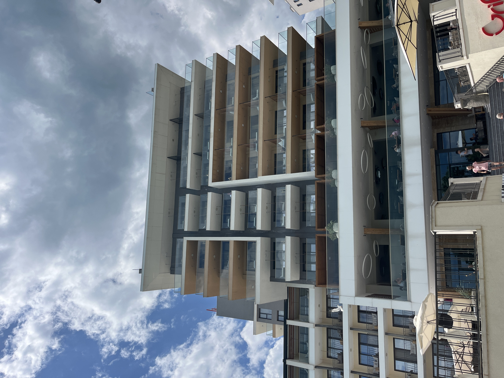 A low-angle shot of a modern building with a facade consisting of glass panels, wooden louvers, and white concrete sections against a cloudy sky. The building is oriented vertically in the frame. The building's right side features a restaurant or cafe with circular design elements and glass partitions. People are visible dining inside the restaurant, while others are walking or standing outside on a patio or balcony. The location is likely in Varna, Bulgaria, due to the architectural style and general urban environment. The sky is partly cloudy, with patches of blue sky visible between the clouds.