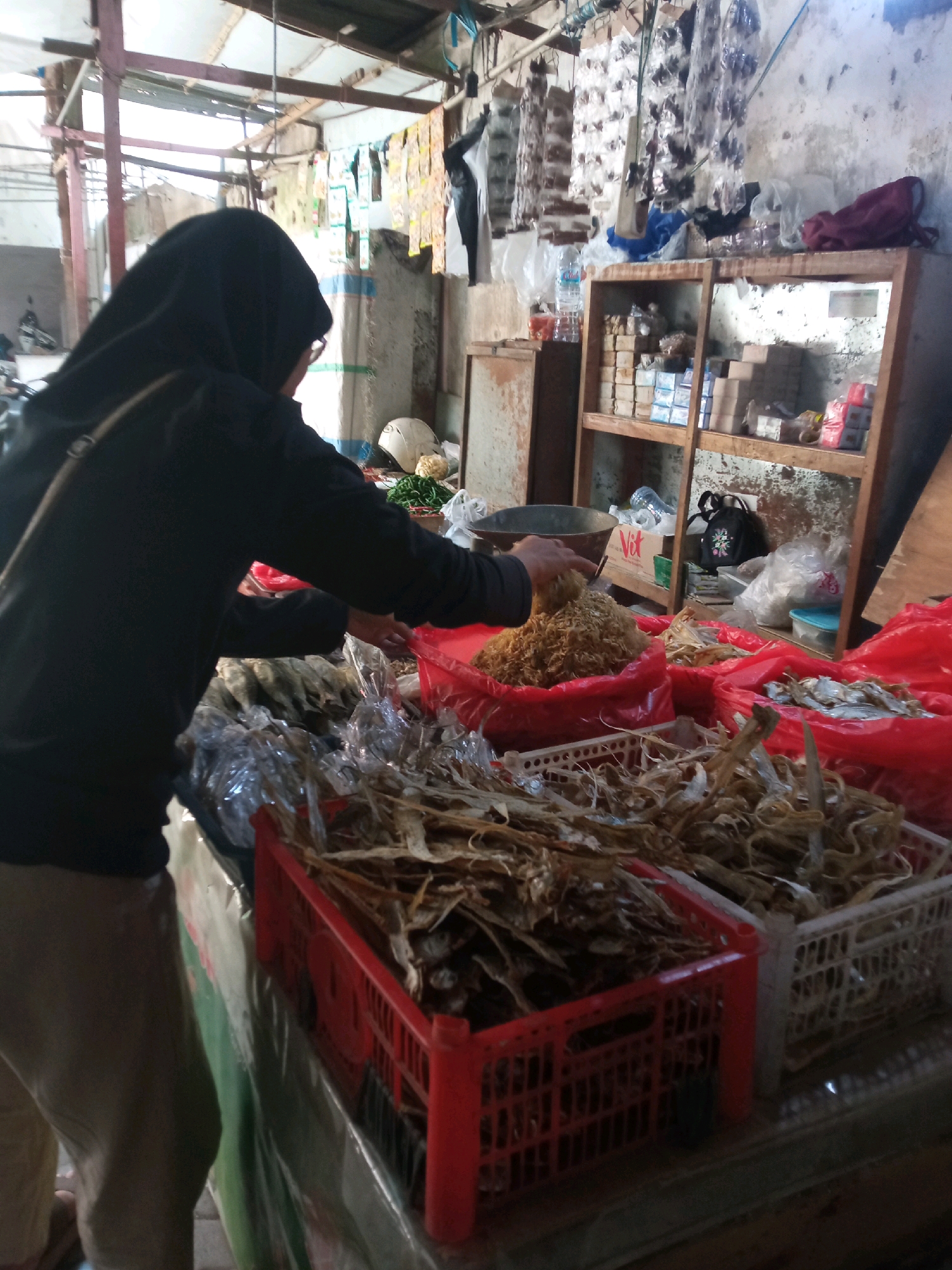 A person wearing a black hijab and a black long-sleeved shirt is standing in front of a stall in an outdoor market in Sambong, Indonesia. The person is using a metal scoop to transfer dried food items, possibly fish or shrimp, from a large red plastic bin to another bin. In front of the person, there are several red and white plastic crates filled with dried food items. In the background, there is a wooden shelf with various items, including bottles of water and packaged goods. The stall appears to be covered by a corrugated metal roof. The overall environment suggests a typical Indonesian market scene.
