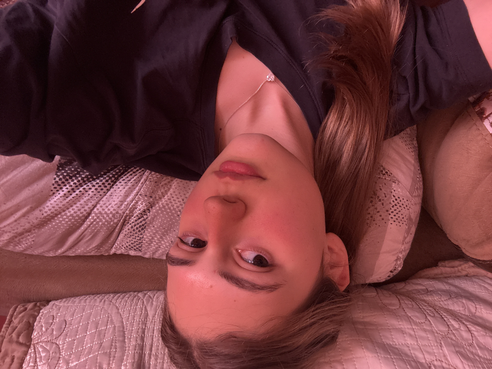 A young woman is lying down, facing the camera. She has fair skin, brown eyes, and long brown hair. She is wearing a dark-colored shirt and a delicate necklace. The background consists of pillows and bedding. There is no information to place the location in Panagyurishte, Bulgaria.