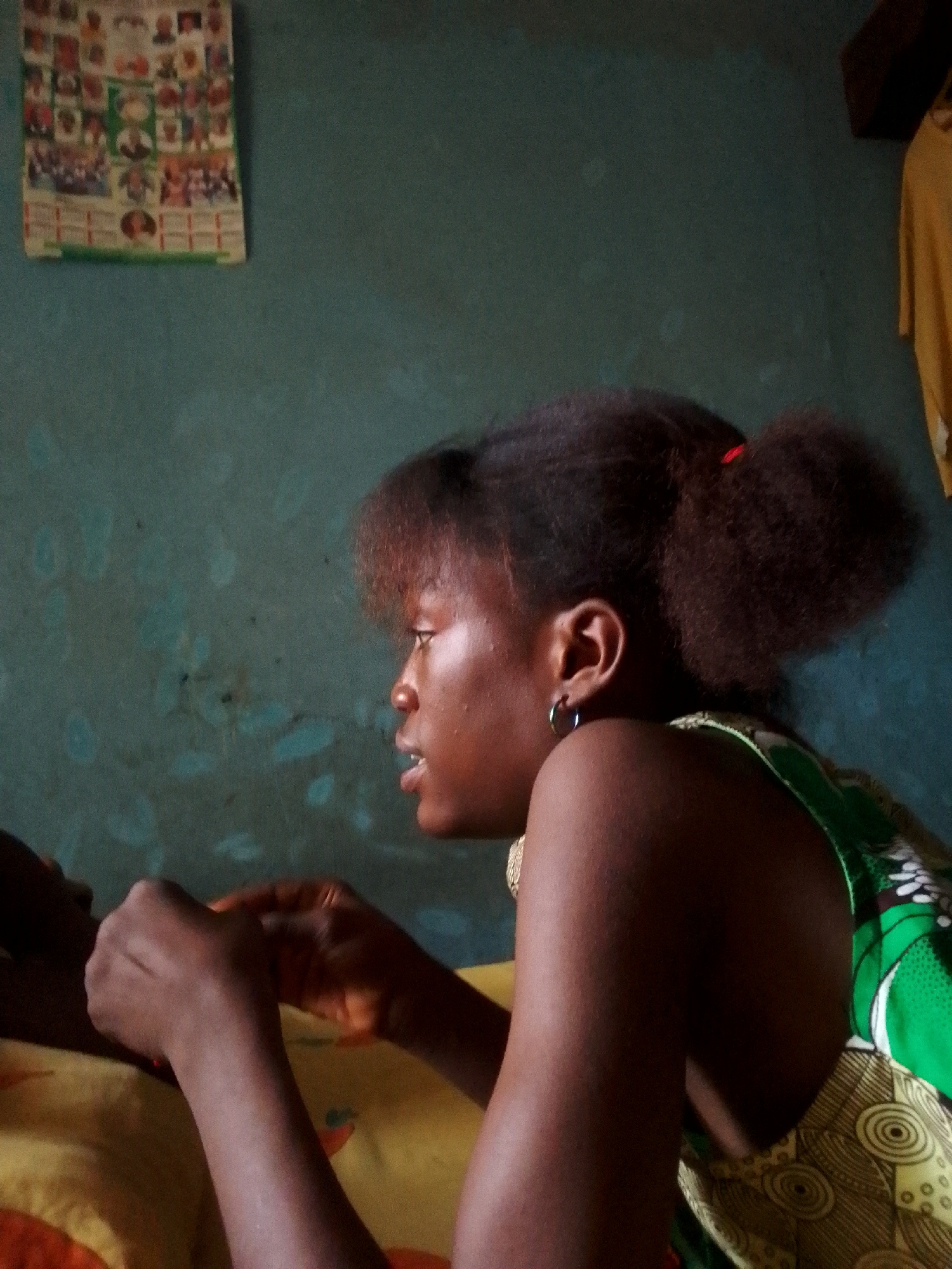 A medium, indoor shot shows a dark-skinned young woman, likely seated or reclining, with her face in profile to the left. Her hair is styled into a high, somewhat puffy ponytail with a red band visible in the back. She is wearing a green and yellow patterned top. Her left hand is slightly raised, and she appears to be looking at something or someone in front of her. Her lips are slightly parted, and she has a small, silver hoop earring in her left ear. The background consists of a blue-green wall with a poster hanging on the upper left and a yellow garment hanging out of focus on the upper right. The specific location is unknown.