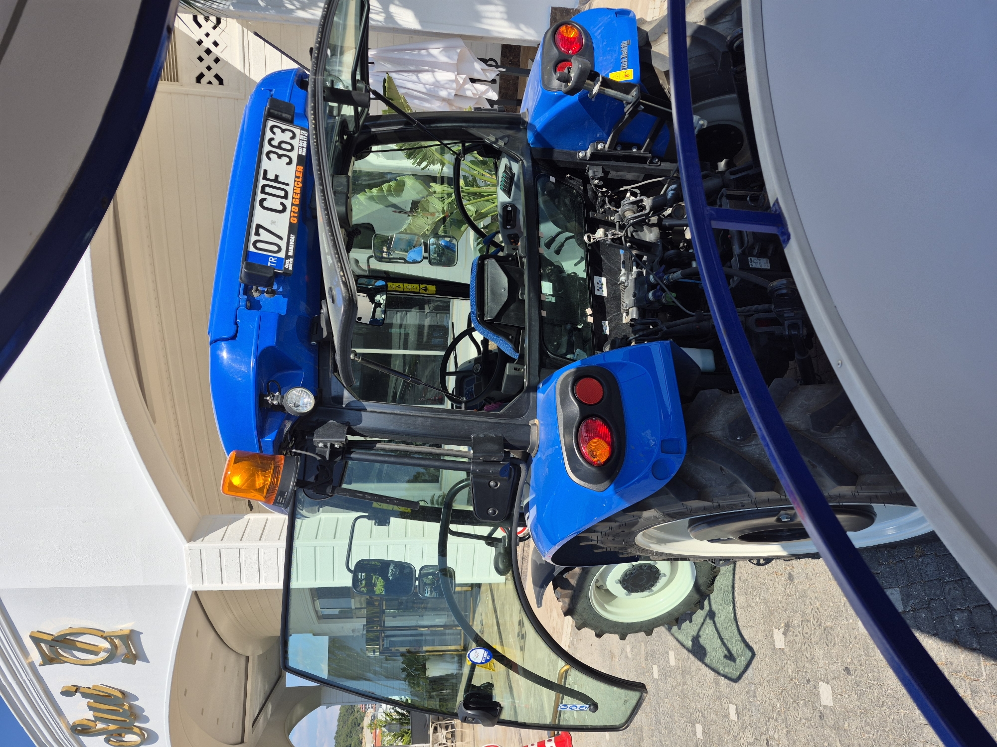 The image shows a bright blue New Holland tractor on display outdoors in Alanya, Turkey. The tractor has the license plate "07 CDF 363." The cab door is open, revealing the interior including the steering wheel. The tractor is positioned in front of a building with a white facade and decorative gold lettering. The sun is shining brightly.