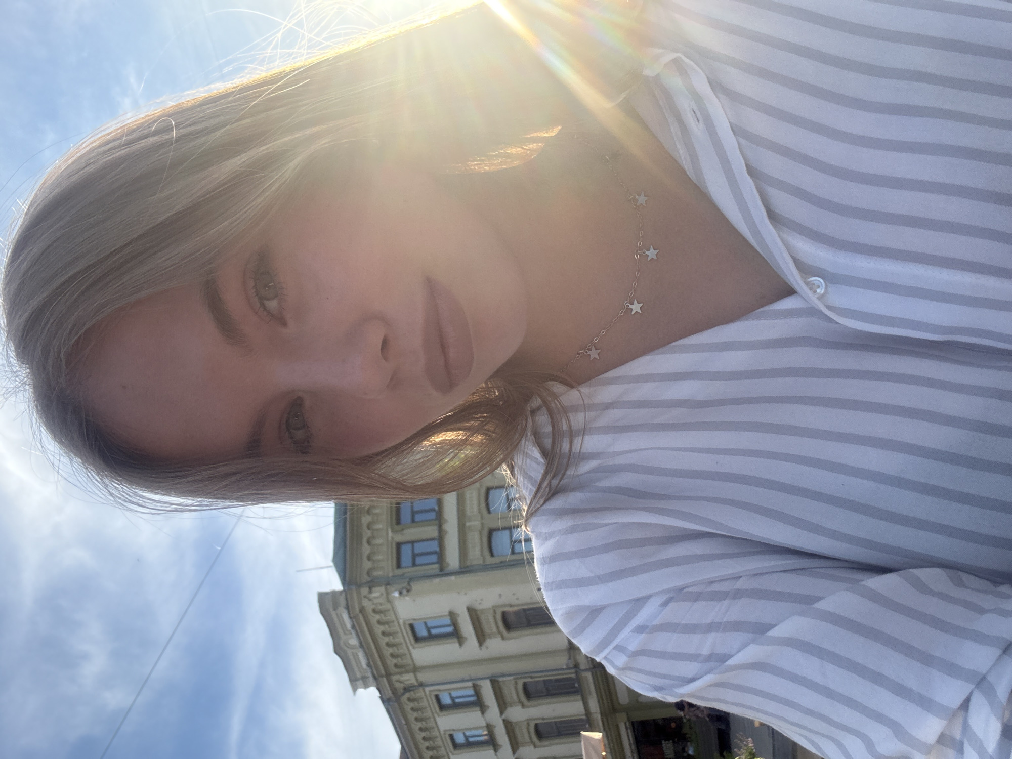 The media file is a selfie of a young woman taken outdoors with a building and the sky in the background. The woman has light brown hair and is wearing a white and grey striped shirt and a delicate necklace with star charms. The sun is shining brightly, creating lens flare and a warm, golden light over her face and hair. Her expression is neutral, and her gaze is directed towards the camera.
The background features a multi-story building with arched windows and architectural detailing, suggesting an urban or historic setting. The sky is a clear blue, with a few wispy clouds. The lighting indicates it is daytime, likely late afternoon or early evening due to the warm, angled sunlight. The image appears to be taken in Nizhny Novgorod, Russia. There is no visible text in the image. The overall impression is a casual and sunny outdoor portrait.