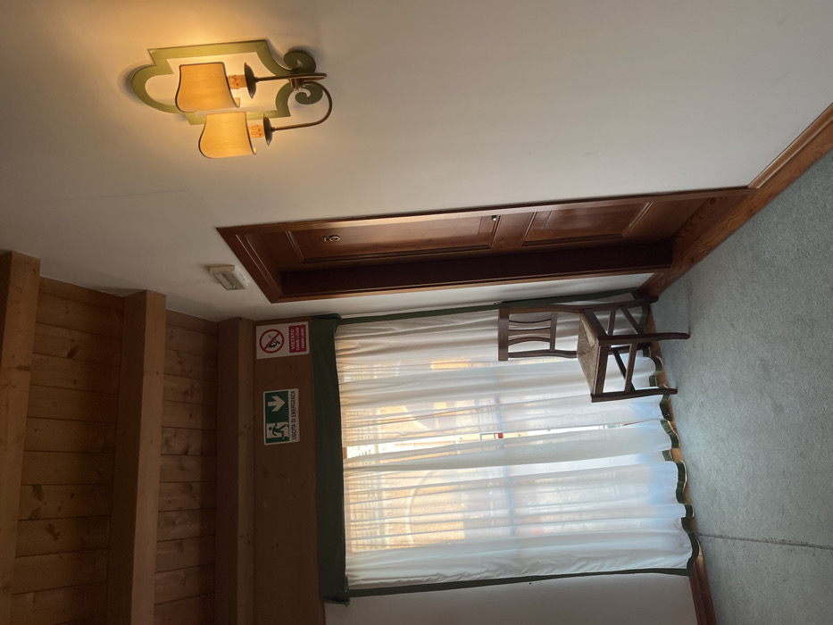 A room features a light fixture on a white ceiling. Wood paneling lines one wall, while a door with a brown frame above a window is adjacent. White curtains with green trim cover the window. An emergency exit sign is on the wall near the window. A wooden chair is positioned in front of the window, and grey carpet covers the floor.