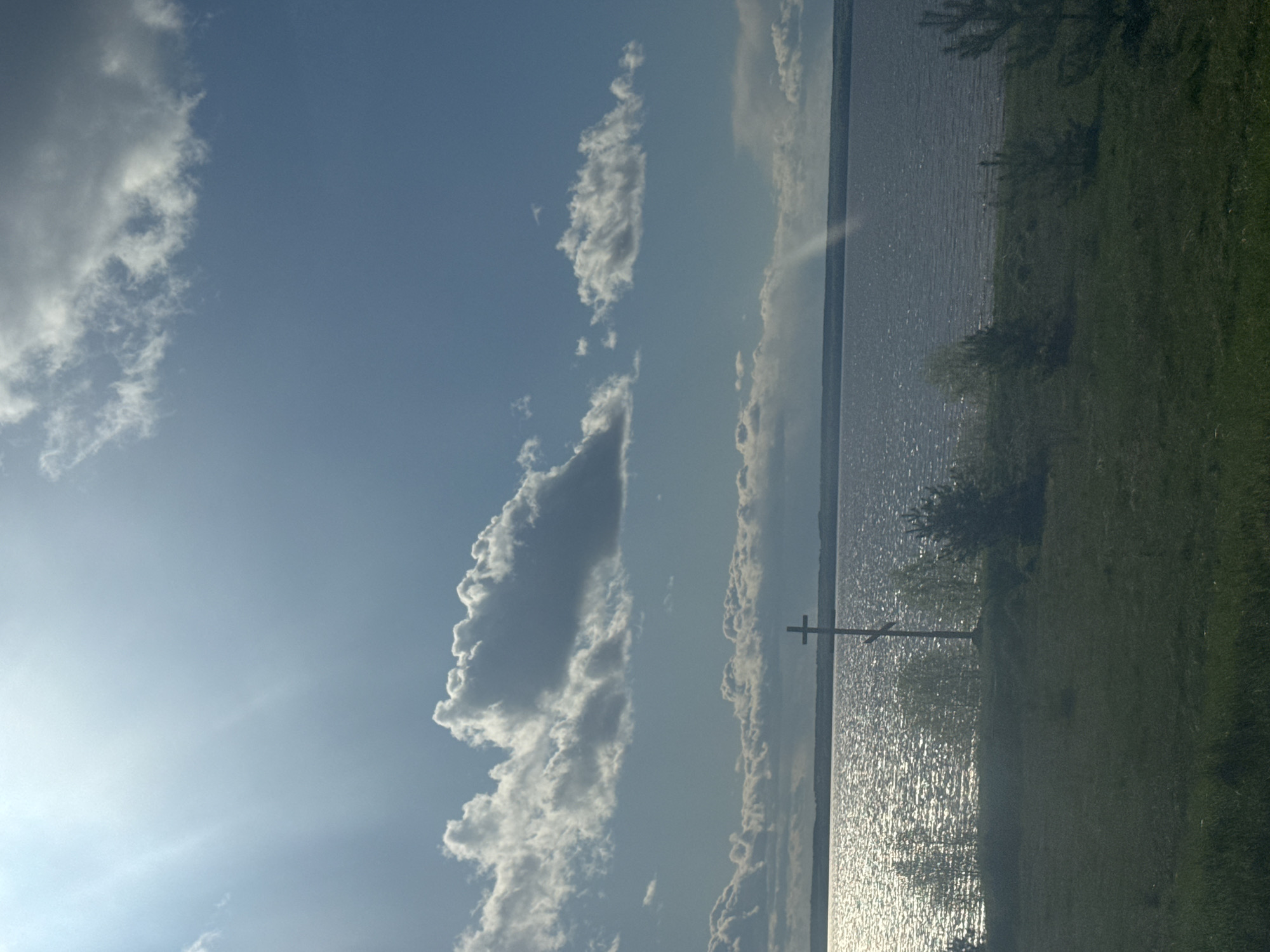 A horizontal image shows a large body of water on the right side, reflecting sunlight, with a cross standing near the shoreline. Adjacent to the water is a green, grassy area. Above the water and land, the left side of the image is dominated by a blue sky with scattered white clouds. The location is suspected to be Kryukovo, Russia.