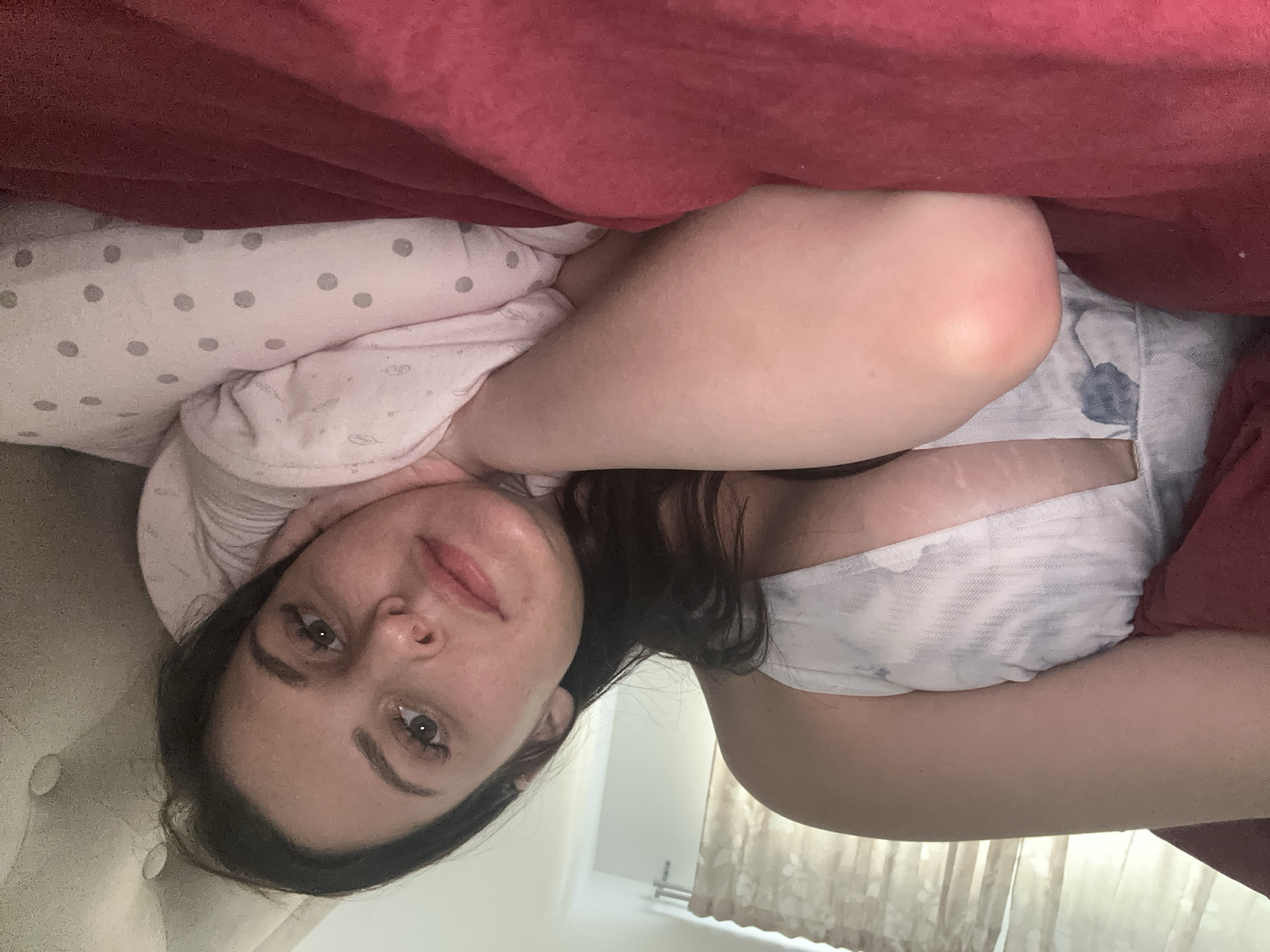 A young woman with dark hair and light skin is lying in bed, propped up on a pillow. She is wearing a white garment with a subtle pattern. She is looking towards the camera with her head resting on the pillow. A pink blanket is visible in the foreground, and a burgundy blanket is partially covering her. The background shows a window with sheer curtains. The overall impression is of a candid, relaxed moment.