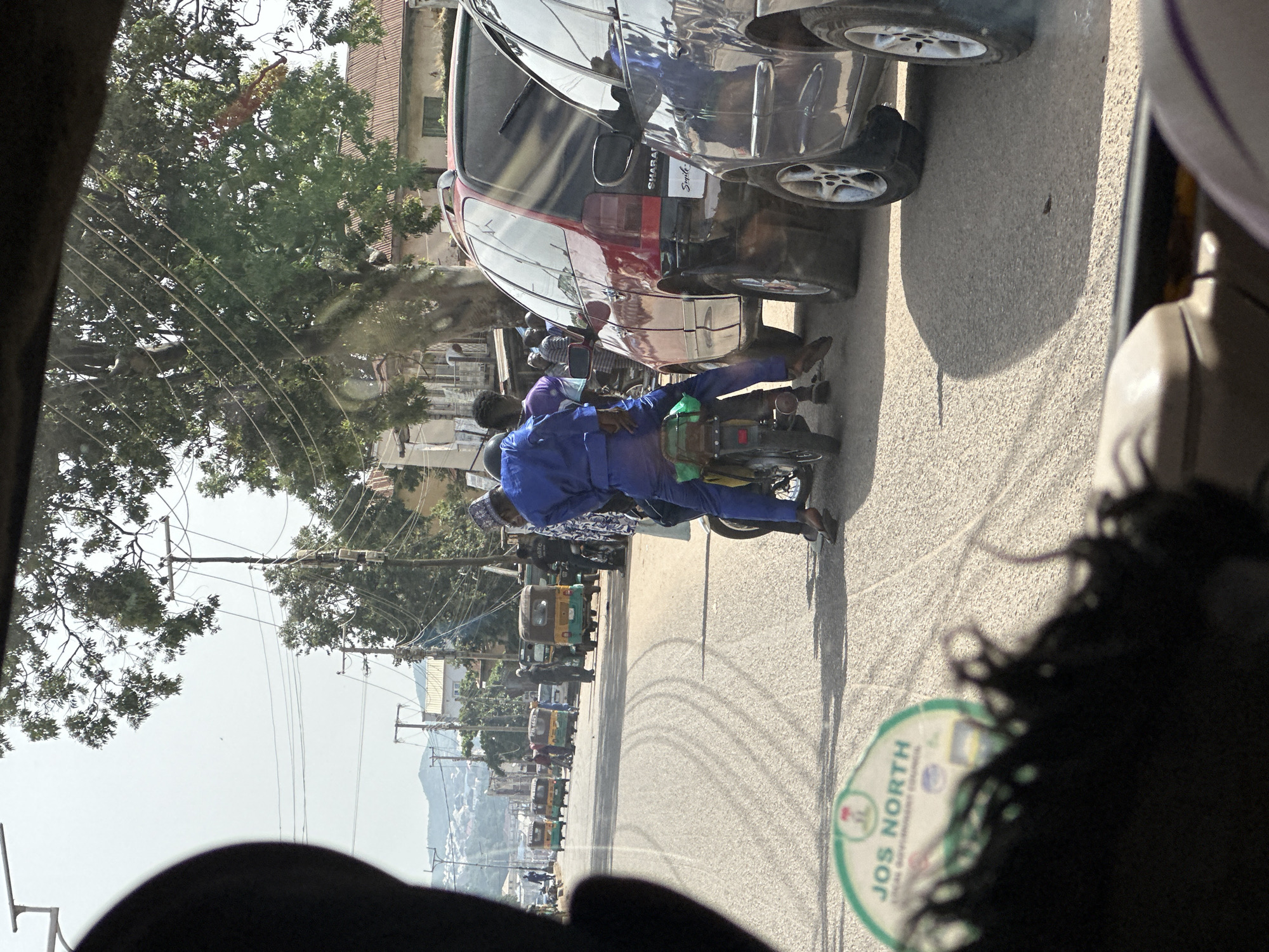 The image, taken from inside a vehicle in Jos, Nigeria, depicts a street scene with several vehicles and people. In the foreground, the top of a dashboard and part of a steering wheel are visible, suggesting the perspective of the photographer. A black fuzzy object, possibly part of the vehicle's interior or a lens cover, is in the lower left corner. A green and white circular sticker with the text "JOS NORTH LOCAL GOVERNMENT COUNCIL" is adhered to the vehicle's surface, indicating the location.
On the road, a motorcycle with two passengers is prominent. The driver, wearing a blue outfit and a cap, is seated at the front. A second person, in a purple top, is seated behind the driver. The motorcycle is moving or stopped in traffic. To the right of the motorcycle, a red SUV is parked or moving slowly. A dark blue sedan is to the right of the red SUV, with its front end obscured.
Further down the street, several auto-rickshaws, commonly known as "Keke Napep" or "Marwa," are visible. They are colored yellow and green. Some are stationary, while others appear to be in motion, suggesting moderate traffic. A few individuals are seen standing near the auto-rickshaws. In the background, buildings with corrugated iron roofs and houses can be seen. Electrical poles and wires are present along the street, and a large tree with dense foliage overhangs the road. Hills and a hazy sky form the backdrop of the scene. The road surface shows tire marks.