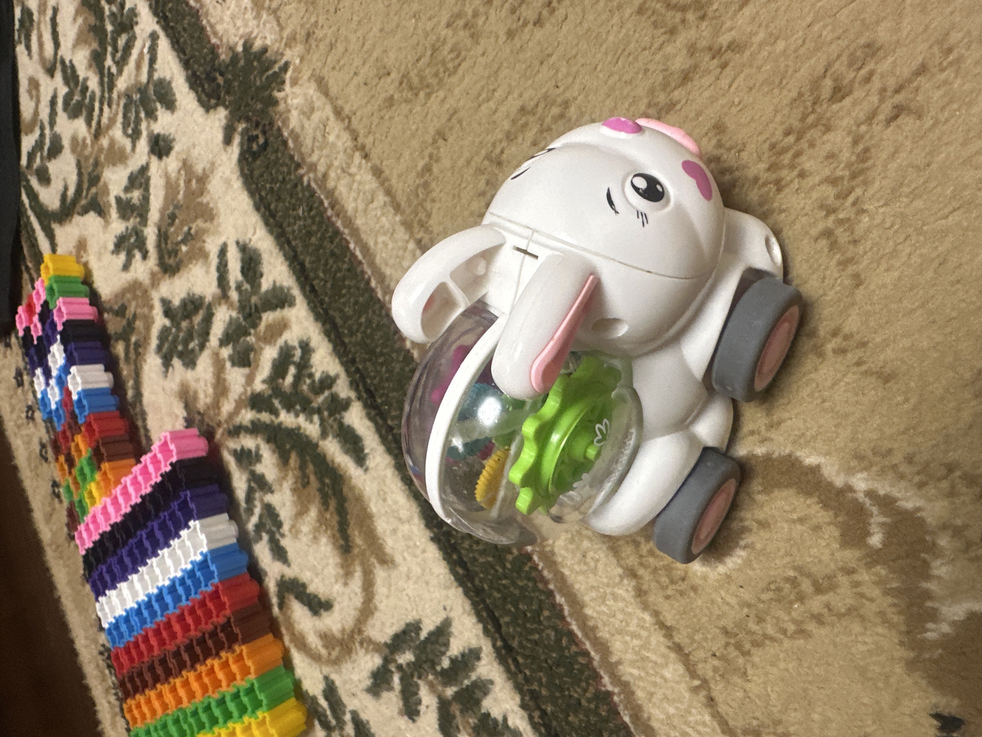 A white plastic toy bunny with pink ears and a pink nose sits on a beige carpet with a floral pattern. The bunny has a clear plastic dome on its back, revealing green and yellow plastic parts inside. To the left of the bunny, a long, colorful string of interlocking plastic blocks is arranged in rows of different colors, including yellow, orange, red, blue, purple, and pink. The scene appears to be indoors.