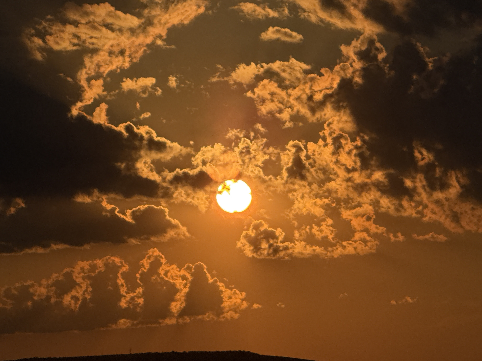 A full shot of a dramatic sunset captures the sun as a bright, glowing orb peeking through a dense arrangement of clouds, casting a warm, orange glow across the sky. The clouds are a mix of dark, silhouetted masses and brightly lit, wispy formations, creating a striking contrast between light and shadow. The sun is positioned slightly off-center to the right, with its brilliant light creating a halo effect around its edges. The scene is framed by a dark silhouette of land in the foreground, suggesting a natural landscape or perhaps the edge of a city at dusk. The overall impression is one of intense natural beauty and a powerful display of atmospheric phenomena.