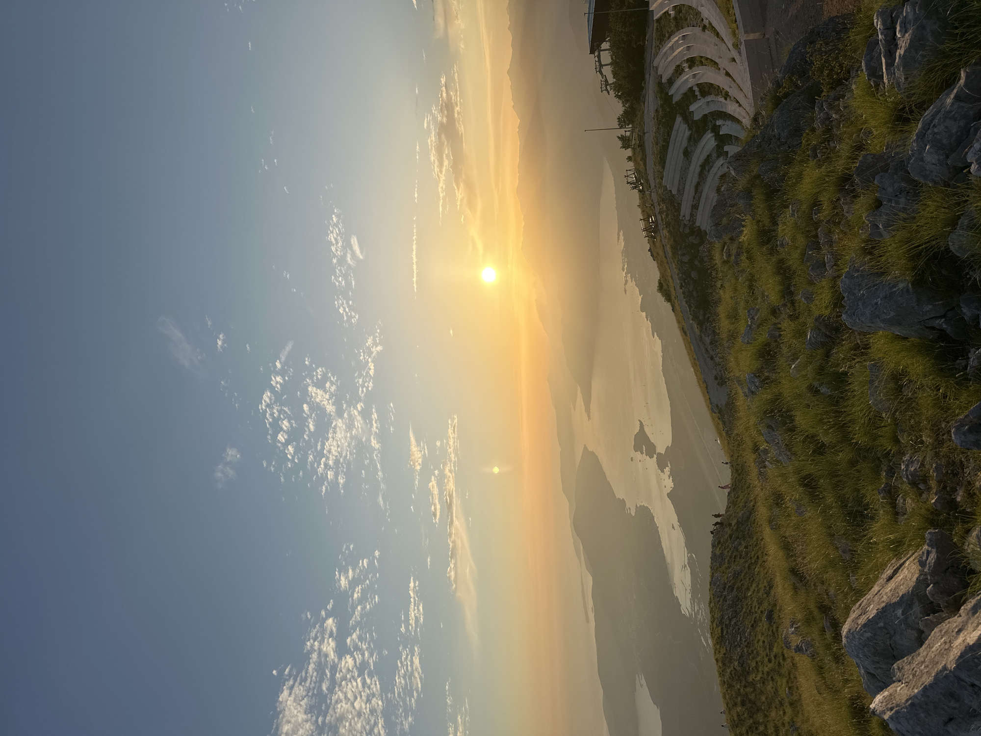 An aerial, wide-angle view captures a sunset over the Bay of Kotor, from a vantage point in Kavač, Montenegro. The sun, a bright orb, hangs low in the sky, casting a warm golden-orange hue across the scene and illuminating scattered clouds. The bay, with its tranquil waters, is nestled between rolling hills and distant mountains, all softened by a gentle haze. On the right side of the image, a tiered outdoor amphitheater is visible, carved into the hillside, with stone seating arranged in curved rows. A pathway leads up to the amphitheater, and a ski lift structure is present near the top of the hill. The foreground is dominated by the rugged texture of a grassy hillside, dotted with large rocks and illuminated by the fading sunlight. Several individuals are scattered across the landscape, some walking along the hillside and others seated in the amphitheater, enjoying the panoramic view. The overall impression is one of serene natural beauty and a peaceful gathering space.