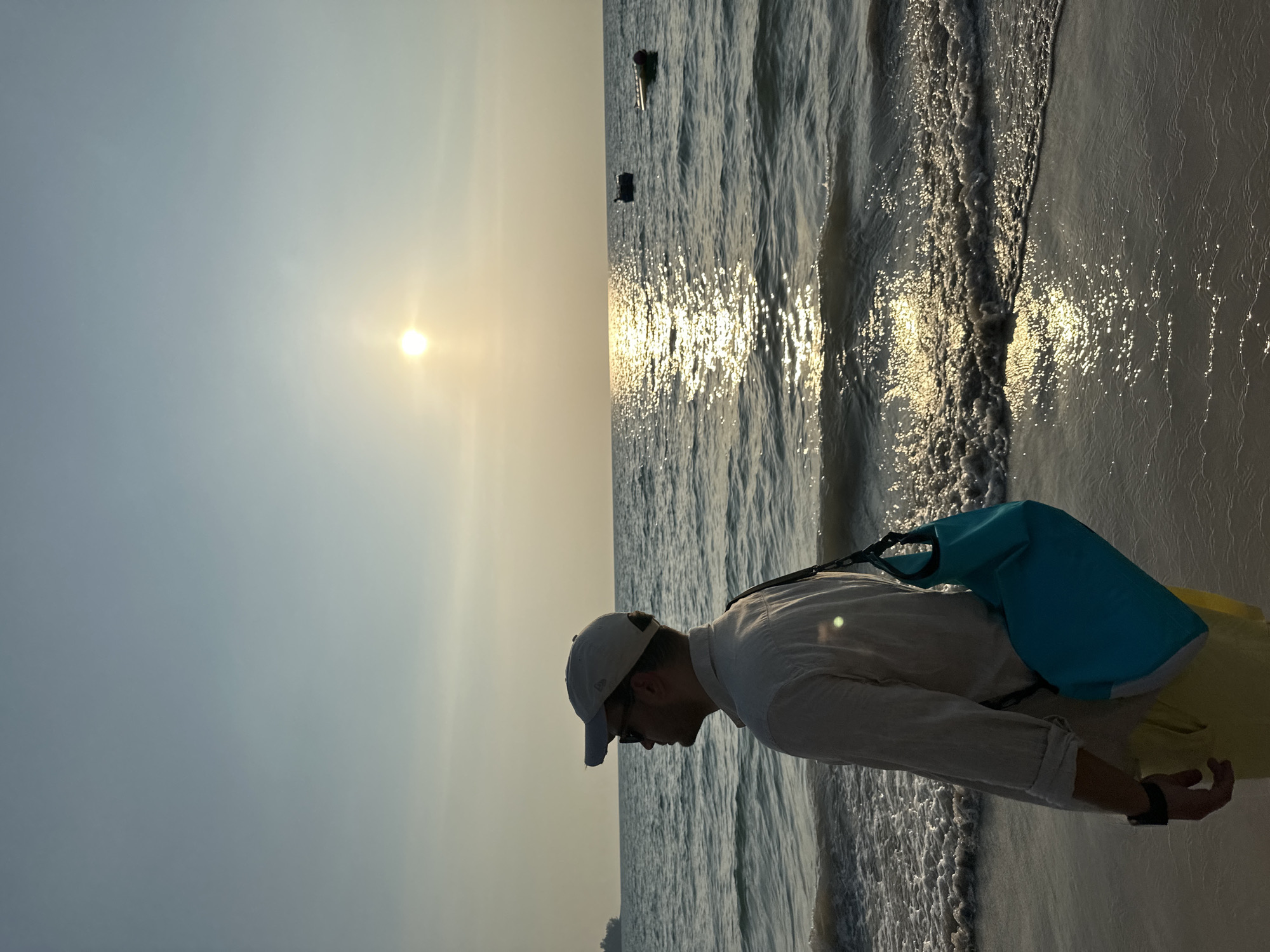 A male individual stands on a sandy beach in Choeng Thale, Thailand, facing left and looking out at the ocean during sunset. He wears a light-colored long-sleeved shirt, yellow shorts, a light-colored baseball cap, and sunglasses. A bright blue cross-body dry bag is slung over his right shoulder. A black watch or wristband is visible on his left wrist. The sun is a bright disc low on the horizon, casting a shimmering path of light across the water. Gentle waves break on the shore. Two small dark objects, possibly boats or buoys, float in the mid-distance on the ocean. A large, dark ship is faintly visible on the far left horizon. The sky is a gradient of light blue and yellow hues.