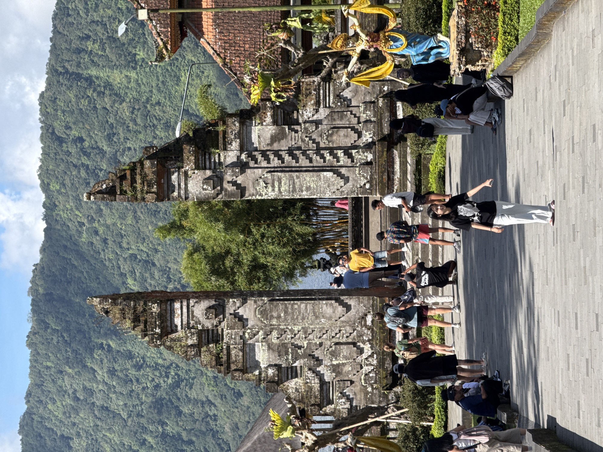 This image depicts the Handara Gate in Baturiti, Indonesia. The gate is a large stone structure with intricate carvings, serving as a popular tourist attraction. Many people are present, some interacting with the gate and posing for photographs, and others walking along the paved path. The background features a lush green mountain landscape and a cloudy sky. Some structures and objects around the gate are decorated in traditional Indonesian styles.