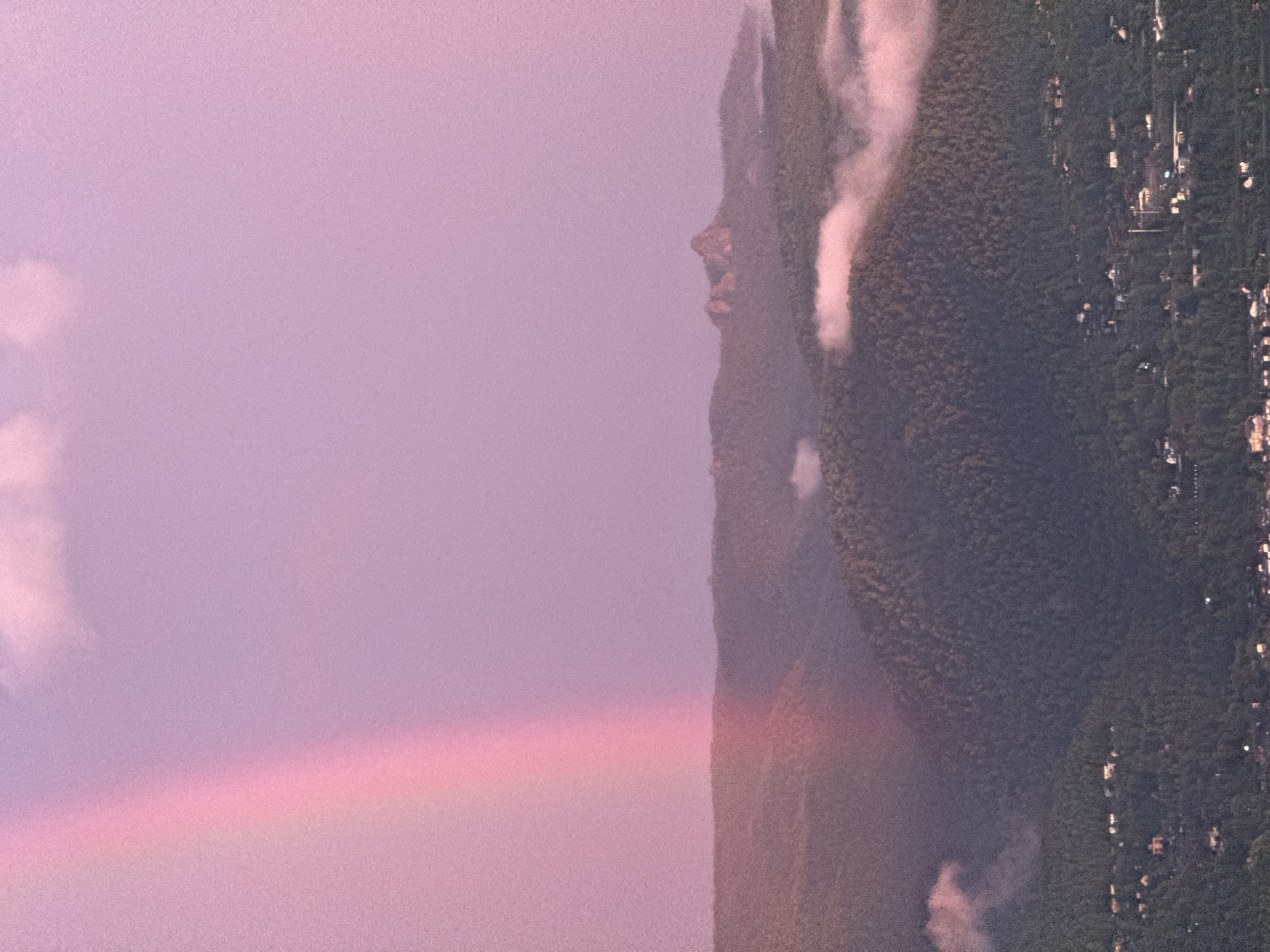 The image depicts a landscape with a prominent pink rainbow arching across a pale purple sky. The foreground features rolling hills covered in dark green trees, with scattered houses visible on the slopes. In the mid-ground, mist or low clouds drift over the tree-covered hills, and a distinctive rock formation, resembling two peaks, rises from a distant hill. The sky transitions from a lighter shade near the top, with hints of white clouds, to a deeper purple towards the horizon. The overall scene is one of natural beauty, possibly captured during or after a rain shower at sunset or sunrise, given the vibrant colors of the rainbow and sky. This view is consistent with scenes from the Stolby Nature Reserve, a protected area located on the northwestern spurs of the Sayan Mountains, near the city of Krasnoyarsk, Russia.