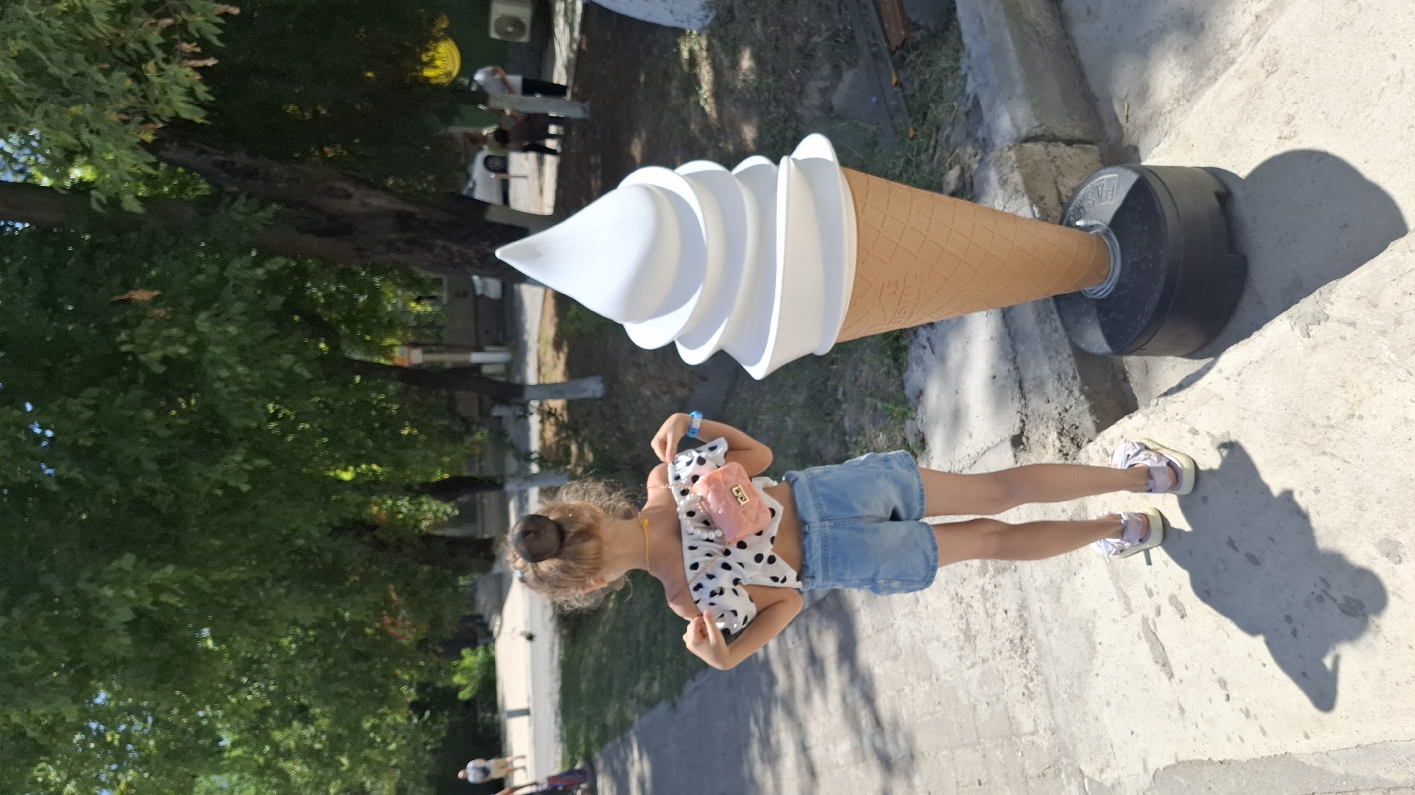 A young girl stands in front of a large, white, soft-serve ice cream cone statue in Shymkent, Kazakhstan. The girl is viewed from behind, wearing a white off-the-shoulder top with black polka dots, denim shorts, and light purple sandals. She has a small pink purse with a chain strap slung over her shoulder. Her hair is pulled up into a bun. The ice cream cone statue is positioned on a black circular base. The scene is outdoors, with trees and a sidewalk visible. The ground is paved with concrete. In the background, there are more trees, a street with a few people and vehicles, and a building. The day is sunny.