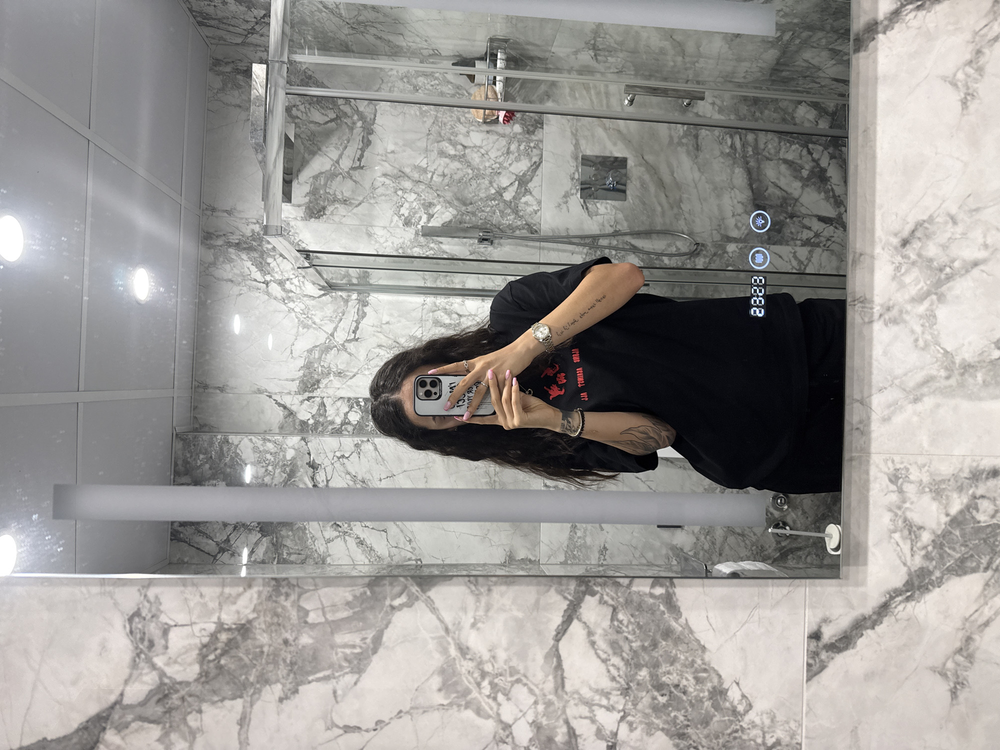 The image shows a mirror selfie taken in a bathroom. The person in the photo, a woman with long, dark, wavy hair and a black t-shirt, is holding a smartphone and taking a picture of herself. The bathroom features marble-patterned walls and a glass shower enclosure. A digital clock displaying "23:23" is visible on the mirror. The environment appears to be a modern and clean bathroom. There is no information provided about the city of Shumen, Bulgaria in the image itself.