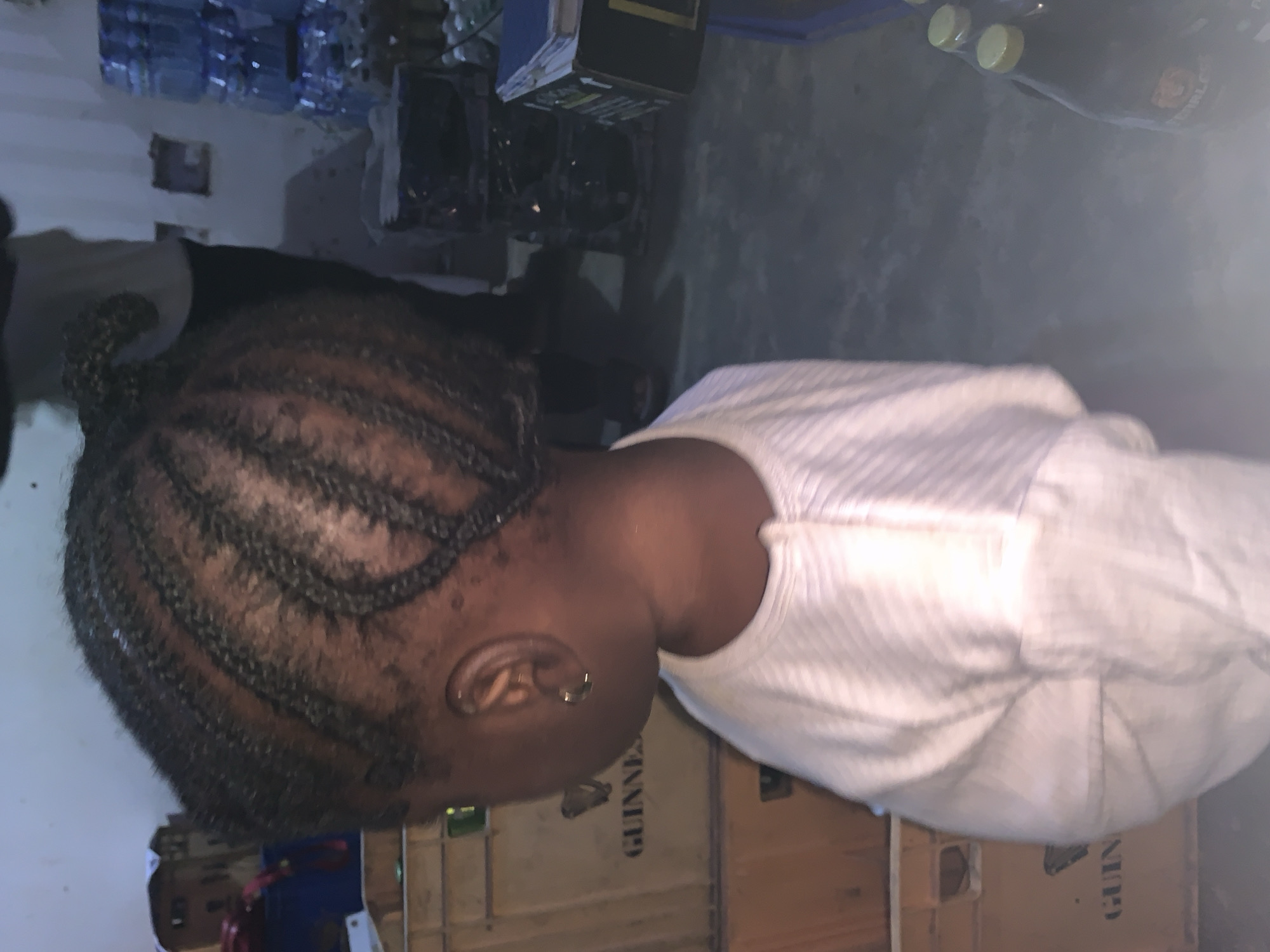 A child with braided hair, wearing a white striped shirt, is positioned in front of a stack of brown Guinness crates. Behind the crates, there's a dimly lit room with stacks of water bottles and other items visible in the background. The photo appears to have been taken indoors. The location provided is Kabong, Nigeria.