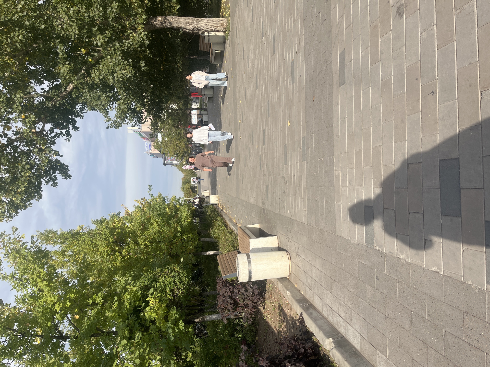 The image depicts a paved pedestrian walkway lined with trees and benches, suggesting a park or public square in a city. In the background, there are buildings with signage, indicating an urban environment. Three women are walking on the path, dressed casually. The walkway is constructed of large, rectangular stone pavers. On the left side of the walkway, there are trees, bushes, a bench, and a cylindrical trash receptacle. The weather appears to be overcast but bright. The scene is captured from a slightly elevated perspective, with the photographer's shadow visible in the foreground. While the provided text mentions Astana, Kazakhstan, this visual information alone does not explicitly confirm the location.