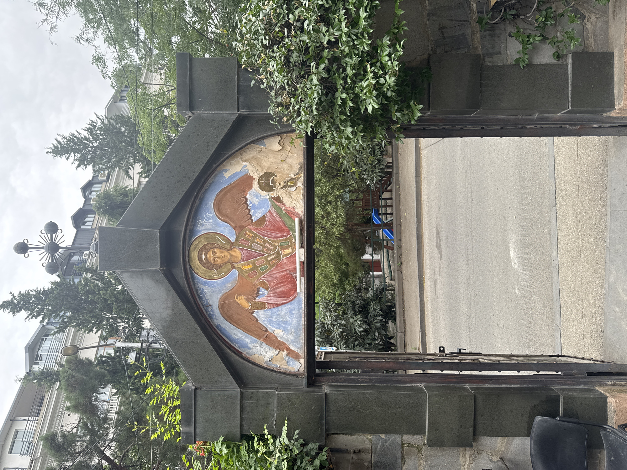 The image shows an arched entryway into what appears to be a religious compound, possibly a church or monastery, in Tbilisi, Georgia. Above the archway is a fresco of an angel with brown wings, dressed in a red robe adorned with colorful geometric patterns. The fresco is partially damaged, with sections of plaster peeling away. The entryway itself is framed by dark stone pillars and a wrought-iron gate, which is open, revealing a street and trees beyond. Lush green foliage climbs over the stone structure on either side of the entrance. In the background, modern buildings and trees are visible under an overcast sky. A street with a sidewalk runs in front of the entryway.