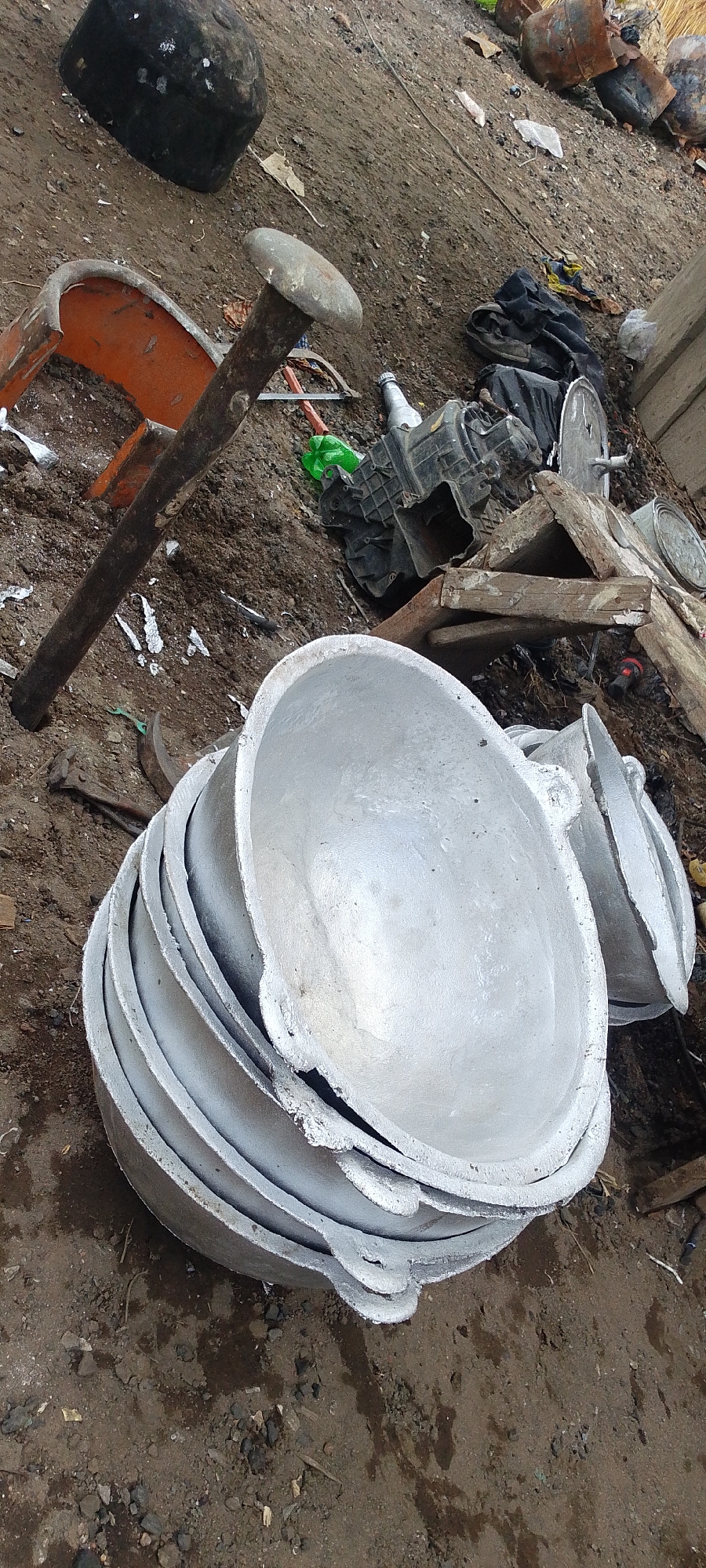 The scene is in an outdoor, possibly industrial, location in Lokoja, Nigeria. The ground is dirt and gravel. A stack of roughly cast, silver-colored metal bowls dominates the foreground. Behind the bowls are various discarded objects, including a broken orange chair, a large metal stake driven into the ground, a black plastic object that could be a machine part, and pieces of wood. In the background, there are black bags or bundles, and other metal items that appear to be scrap or discarded. The scene suggests a junkyard or a recycling area. No people are visible.