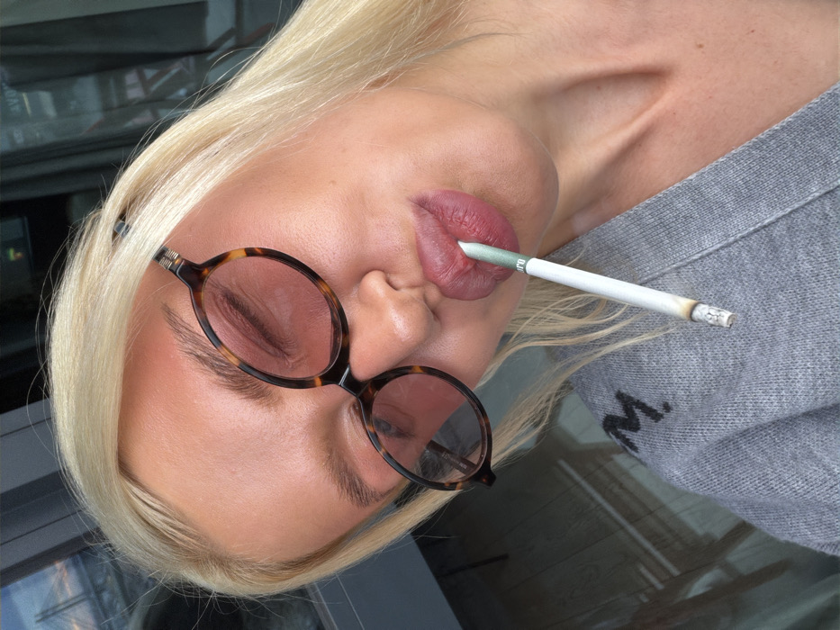 A woman with blonde hair and sunglasses tilts her head while holding a lit cigarette in her mouth. The cigarette has a white filter with a green band and is actively burning. She is wearing a gray sweater with a blurred black letter "M" on it. The background includes a window or reflective surface, indistinctly showing objects and light.