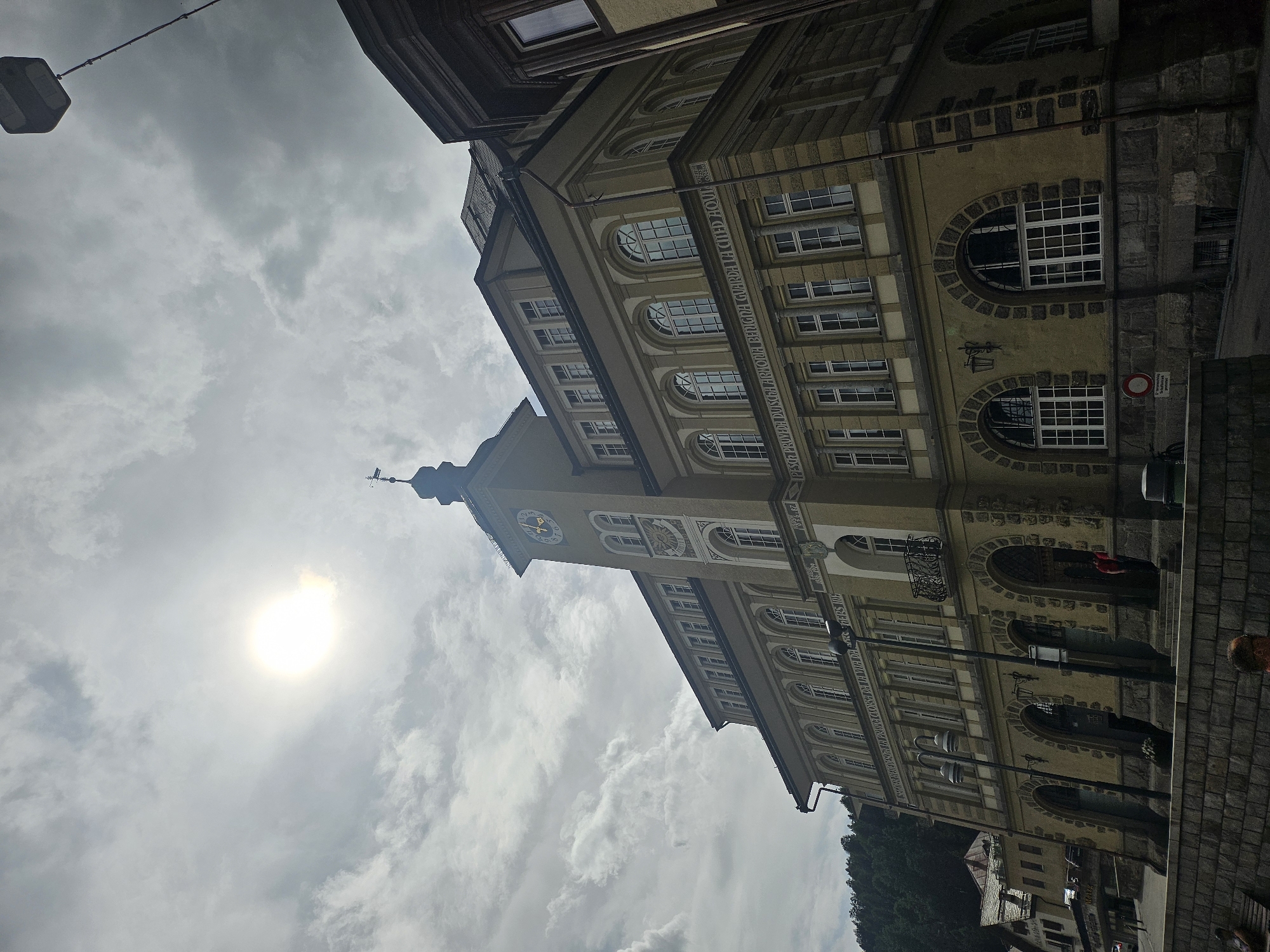 This media file captures an upward-angled view of an old building with a prominent clock tower. The building, with its pale yellow facade and arched windows, appears to be situated in a European town. The clock on the tower shows the time as approximately 3:50. The sky is overcast with dramatic, textured clouds, and the sun is visible as a bright, hazy orb, suggesting either early morning or late afternoon. The scene is one of a historical urban environment, possibly in St. Moritz, Switzerland, given the context. There is a street with cobblestones in the foreground and the architecture suggests a place with a rich history. The presence of streetlights and what appears to be a traffic sign with German text, reading "Vorsicht Rutschig," which translates to "Caution Slippery," further hints at a European location. A few indistinct figures are visible on the street, and a person with red clothing is seen in an arched doorway, but their activities are not clearly discernible. The overall mood is somewhat dramatic due to the sky and the angled perspective.