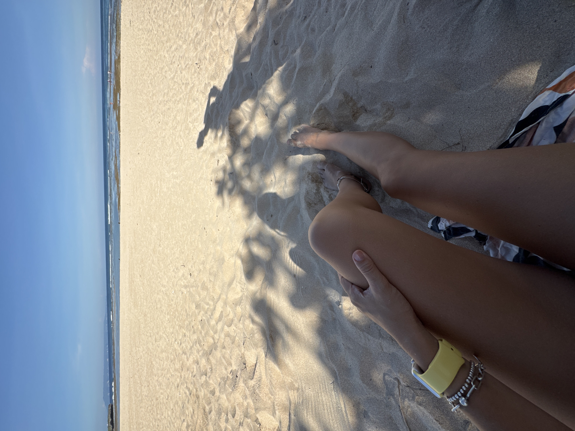 The photograph depicts a person relaxing on the sand at a beach in Kuta Selatan, Indonesia. The beach stretches along the coastline, meeting the ocean under a clear blue sky. A person is positioned in the foreground, reclining on the sand. One hand is visible, wearing a yellow smartwatch and several bracelets. The individual's bare legs stretch out onto the sand, with toes visible at the top of the image. The shadow of a tree or foliage is cast upon the sand in the central part of the picture. The sand is light tan in color. The edges of what appear to be a piece of clothing, potentially a swimsuit or beach towel, are seen along the upper right side of the frame.